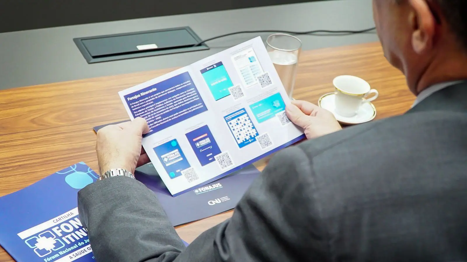 Vista em ângulo superior e por trás de um homem sentado à mesa, segurando um folheto aberto do 