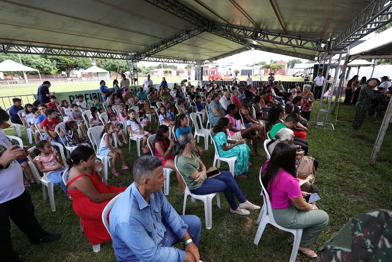 Público diverso sentado em cadeiras brancas sob uma grande tenda aberta em gramado. Ao fundo, árvores e uma ambulância. O ambiente é de um evento comunitário e institucional ao ar livre.