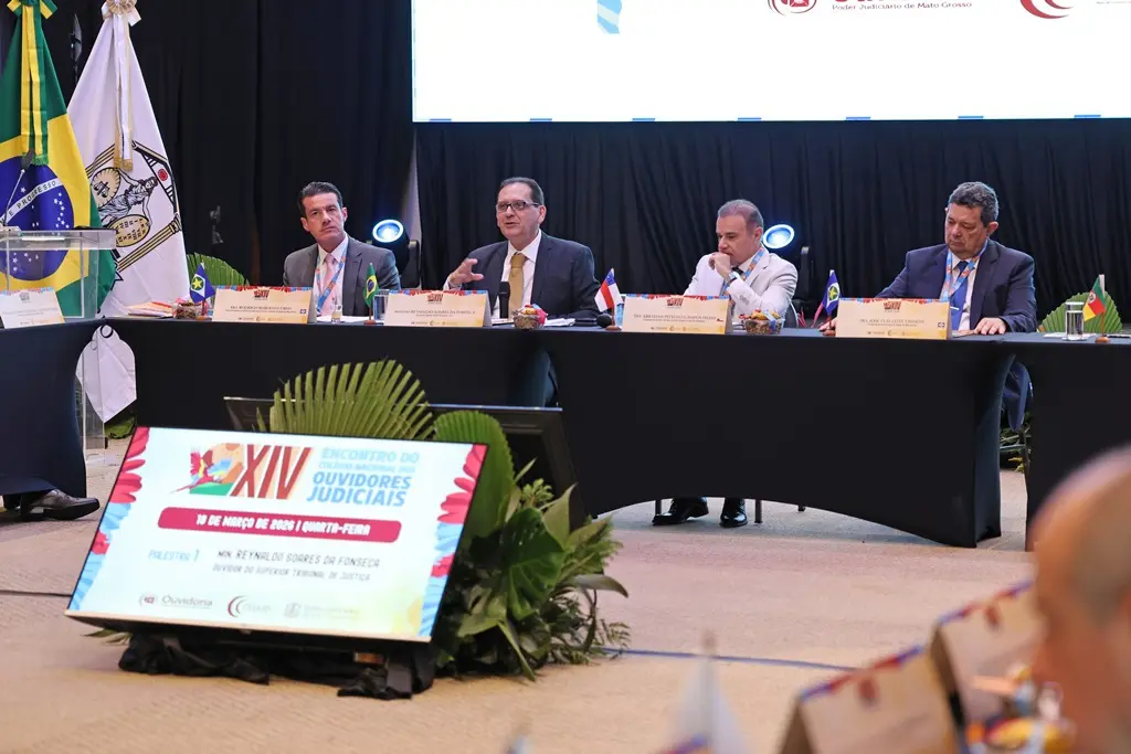 Foto horizontal em plano aberto que mostra o dispositivo de honra do Encontro do Colégio Nacional de Ouvidores Judiciais, composto pelo ministro do STJ, Reynaldo da Fonseca, que fala ao microfone, ladeado pelo presidente e pelo vice-presidente do Cojud.