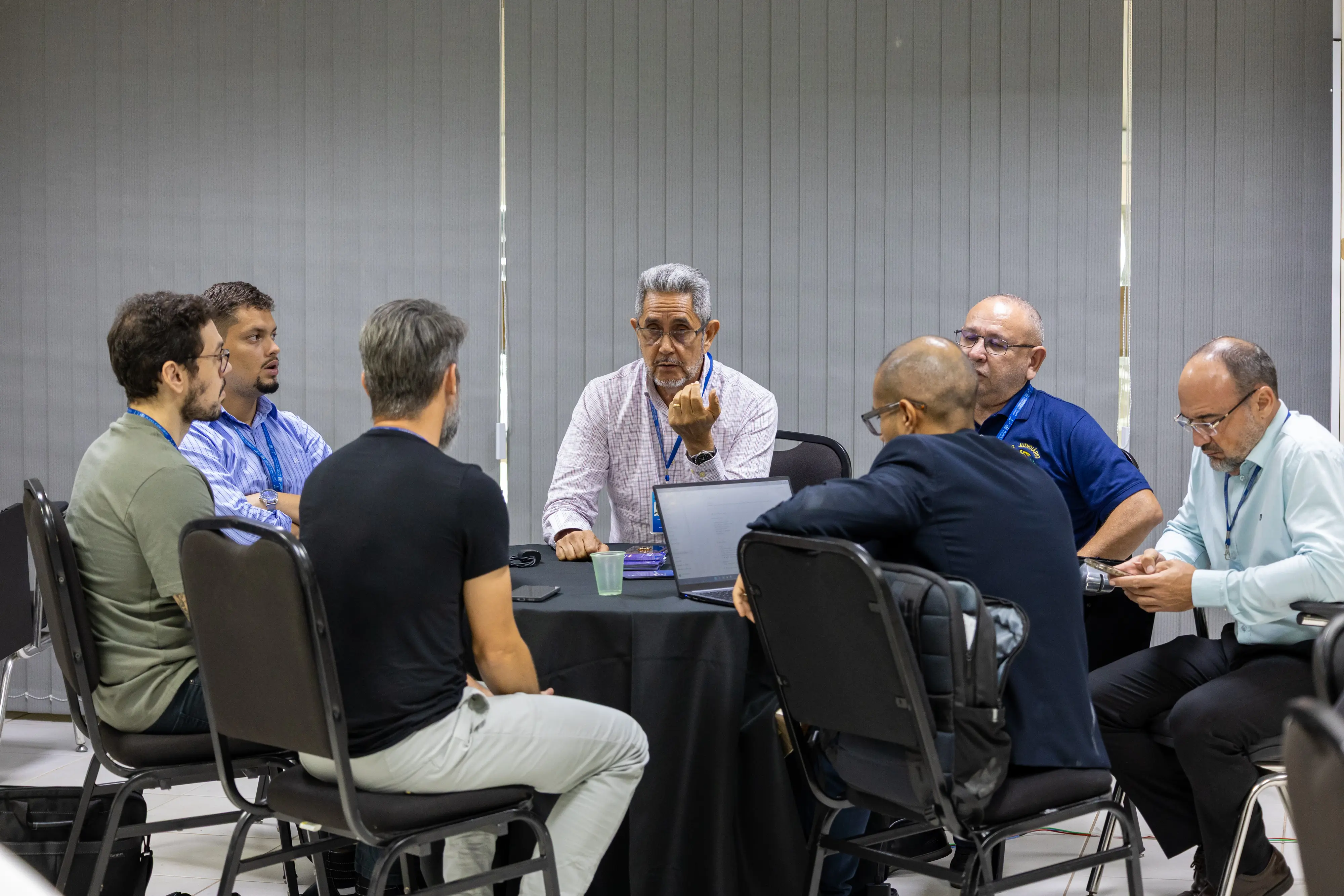 Grupo masculino reunido em círculo debate proposta. Homem ao centro gesticula ao falar, liderando a conversa. Notebook aberto e celulares apoiam a discussão colaborativa durante a oficina.