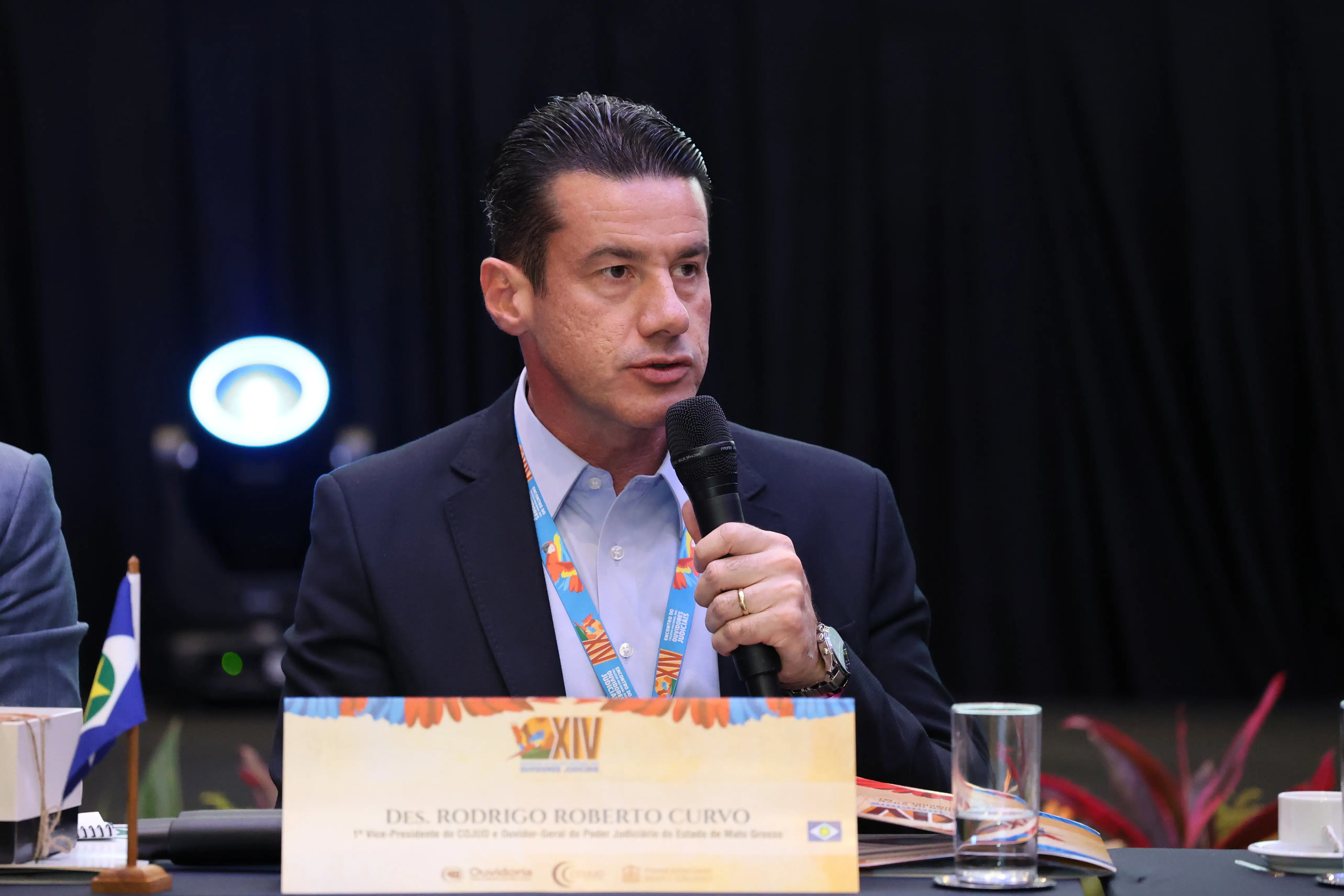 Homem de terno escuro fala ao microfone durante reunião. Usa crachá do evento e está sentado à mesa com identificação nominal à frente, além de copo d’água e materiais de apoio. Expressão firme e atenta.