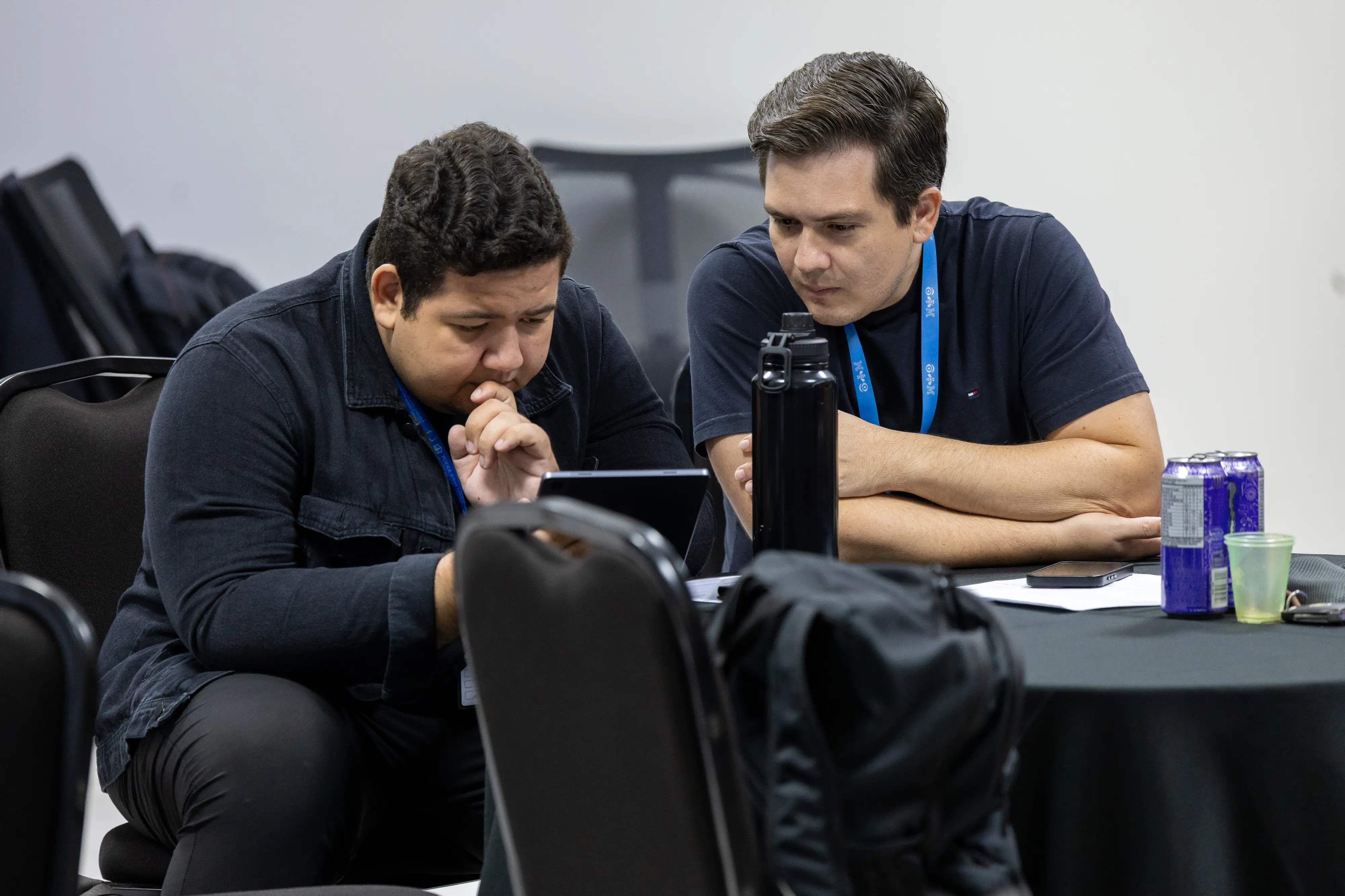 Dois homens analisam conteúdo em tablet apoiado na mesa. Um mantém a mão no queixo, concentrado, enquanto o outro observa atentamente. Garrafa térmica e latas indicam jornada de trabalho prolongada.