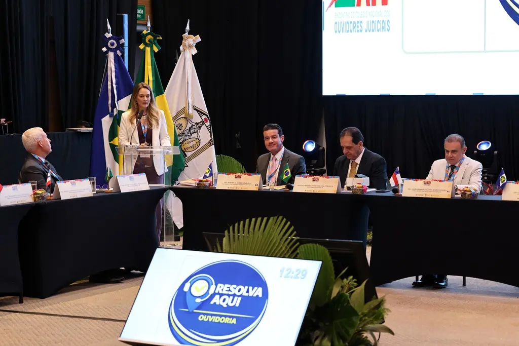 Foto horizontal em plano aberto que mostra a coordenadora da Ouvidoria do Tribunal de Justiça de Roraima, Tainah Westin, falando no púlpito para o público. No dispositivo de honra, estão desembargadores e o ministro do STJ, Reynaldo da Fonseca.