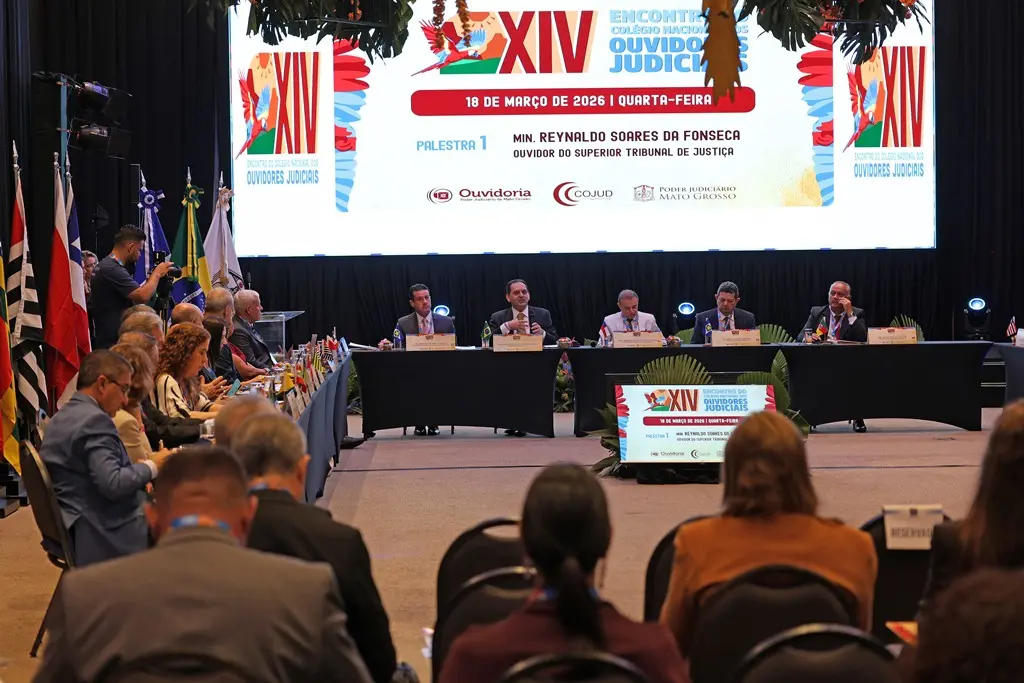 Foto horizontal em plano aberto que mostra o salão onde ocorreu o Encontro do Colégio Nacional de Ouvidores Judiciais, com um grande telão com a logomarca do evento, integrantes do colégio sentados no dispositivo de honra e participantes sentados na plateia. 