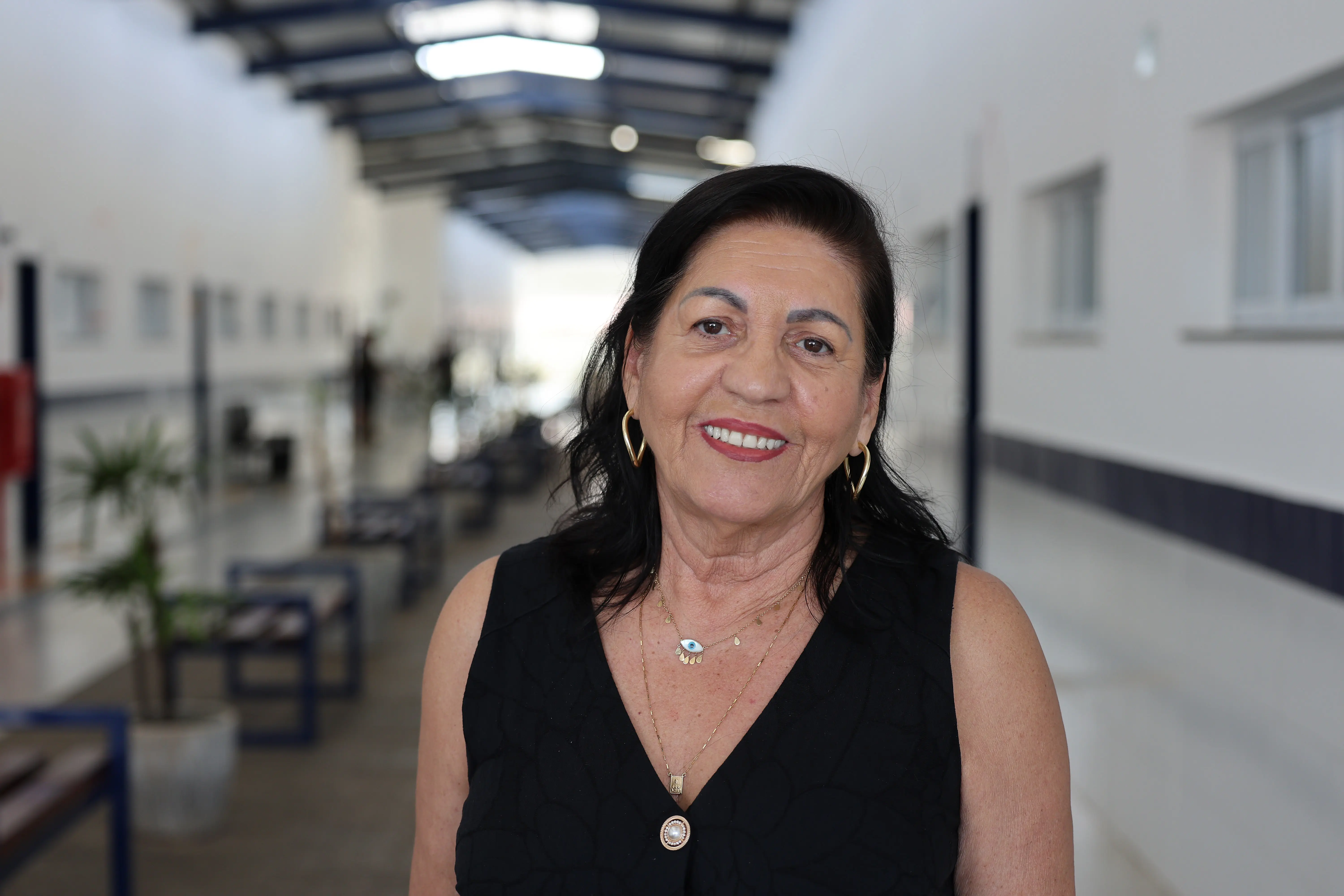 Retrato de Maria Denise de Souza Carvalho. Mulher de pele clara, cabelos pretos e sorriso gentil. Ela usa brincos dourados e colares. O fundo é um corredor escolar iluminado e levemente desfocado.