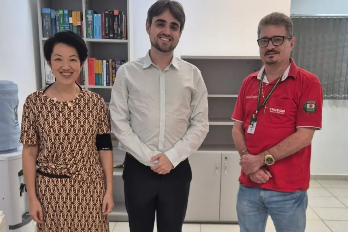 Uma mulher com vestido estampado e dois homens — um de camisa social branca e outro de polo vermelha do Procon — posam sorridentes em frente a uma estante de livros jurídica.