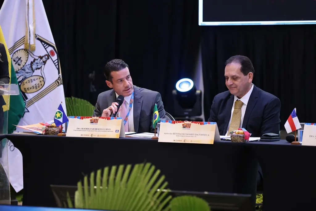 Foto horizontal que mostra o desembargador Rodrigo Curvo sentado ao lado do ministro ouvidor do STJ, Reynaldo Soares da Fonseca, falando ao microfone e voltado para o ministro. 