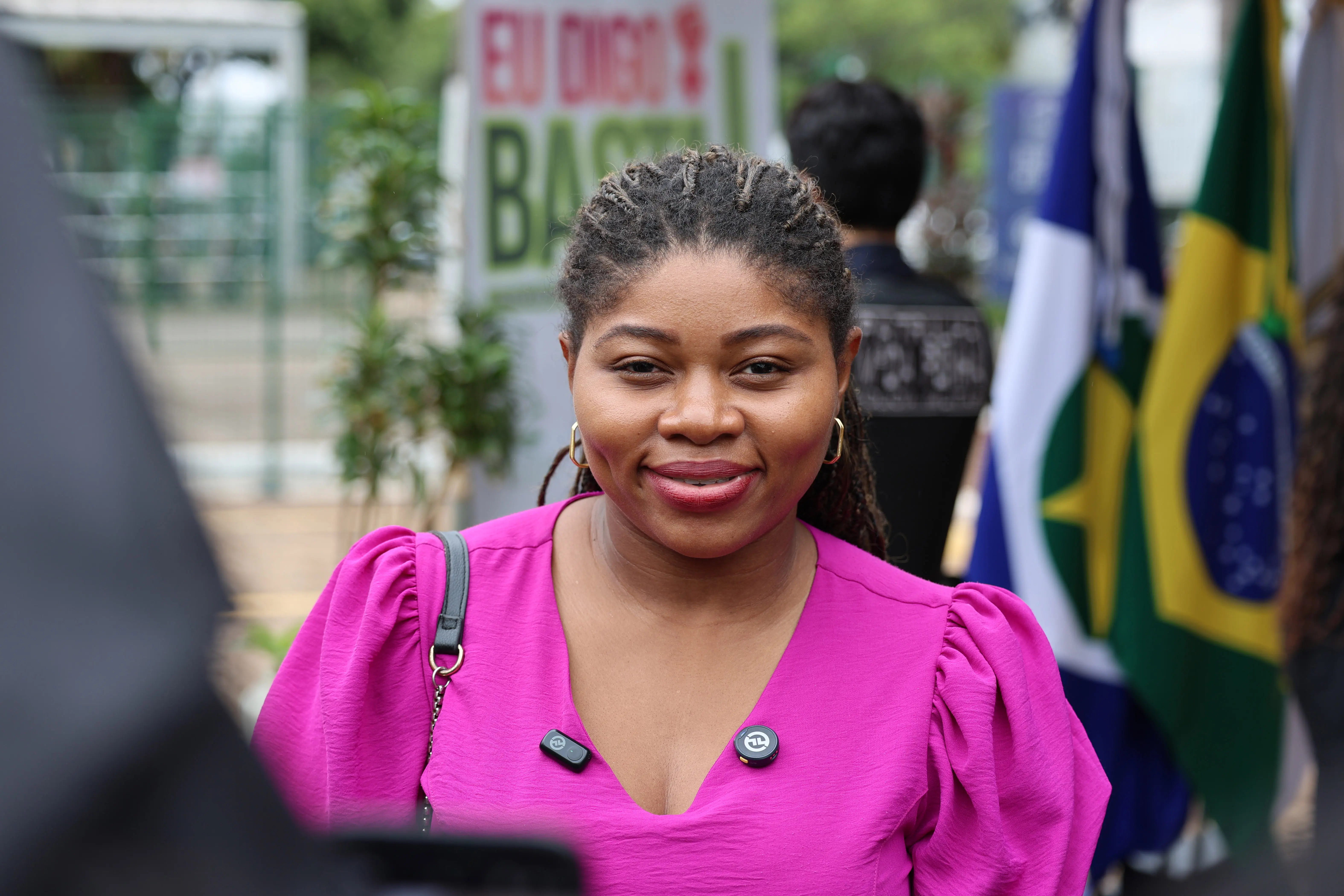 Josiane Delfina Leite, mulher negra com cabelos em tranças raiz. Sorri frontalmente usando blusa rosa de mangas bufantes e decote em V. Ao fundo, banner da campanha e a bandeira do Brasil.