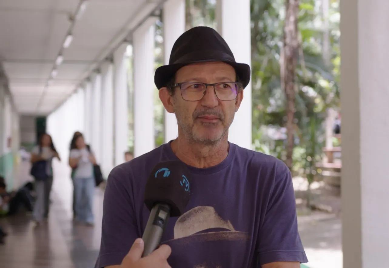 Foto horizontal em plano médio que mostra o professor Luiz Renato de Souza durante entrevista à TV Justiça, no corredor do IFMT. Ele é um homem pardo, alto, magro, usando camiseta azul estampada, chapéu preto e óculos de grau.