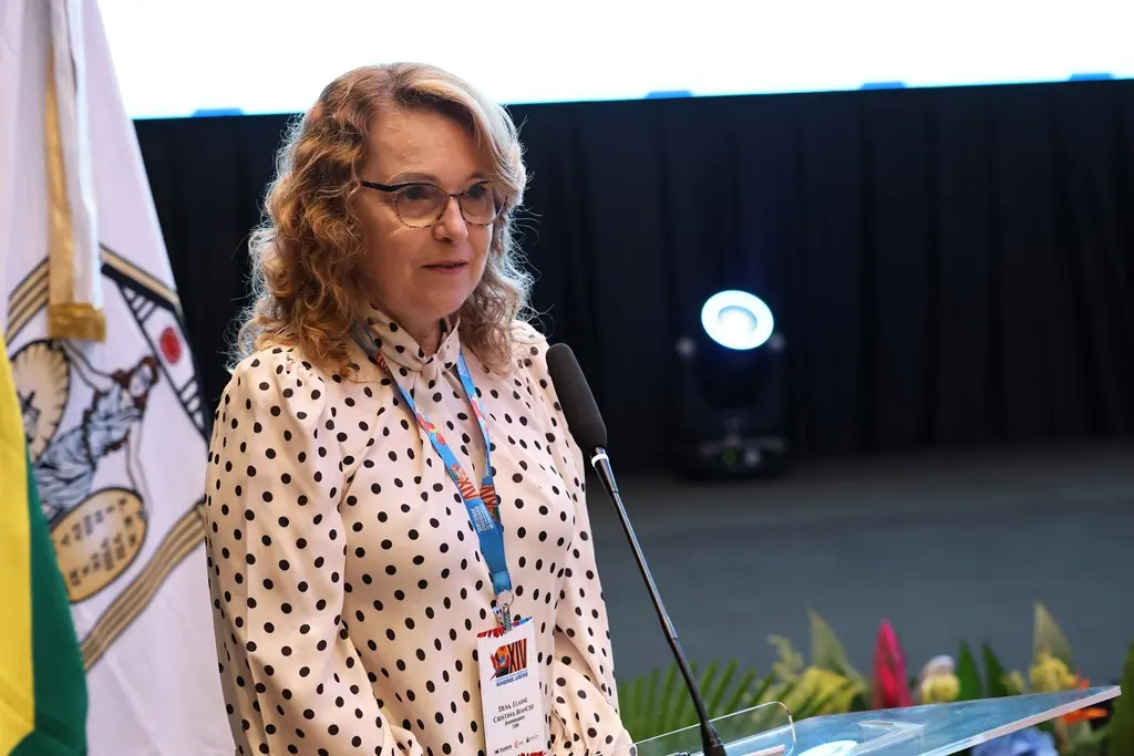 Foto horizontal que mostra a desembargadora do Tribunal de Justiça de Roraima, Elaine Bianchi, falando no púlpito. Ela é uma mulher branca, de cabelos loiros e cacheados, usando blusa de manga comprida branca de bolinhas pretas, óculos de grau e crachá do Cojud.
