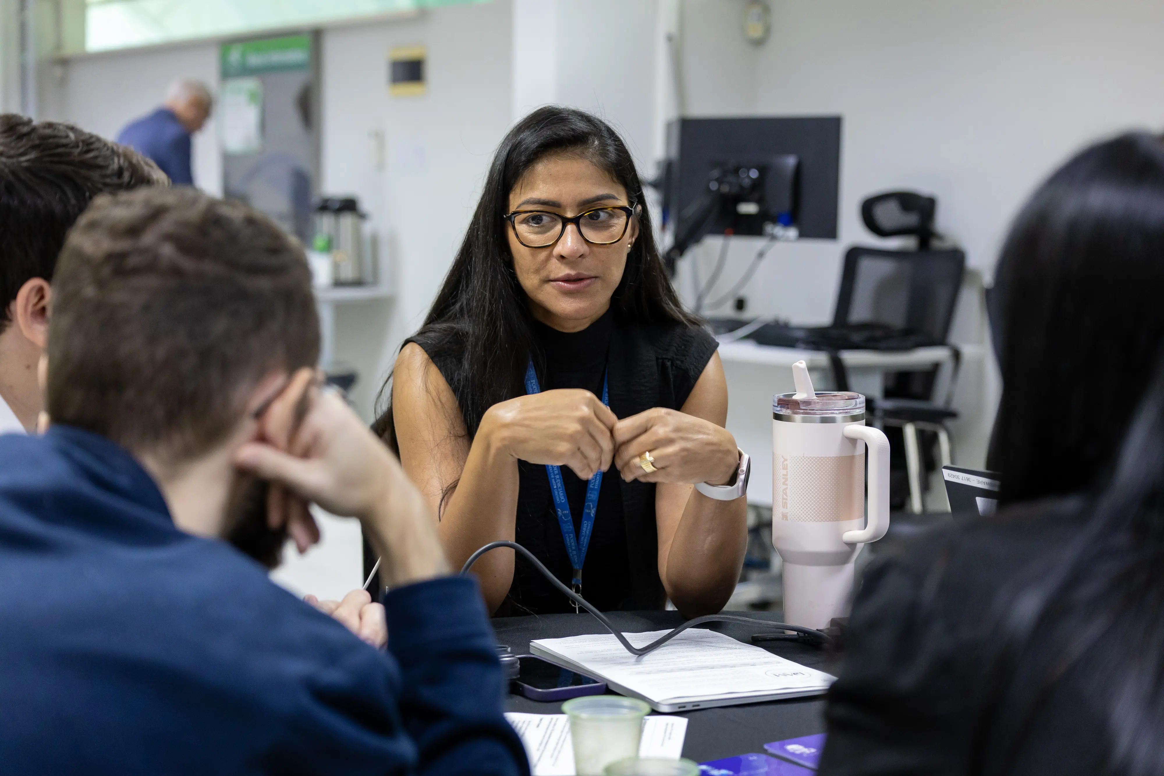 Mulher de óculos conduz diálogo em mesa redonda. Com as mãos entrelaçadas, explica ponto aos colegas atentos. Sobre a mesa, celular, cabos e copo térmico acompanham documentos da atividade.