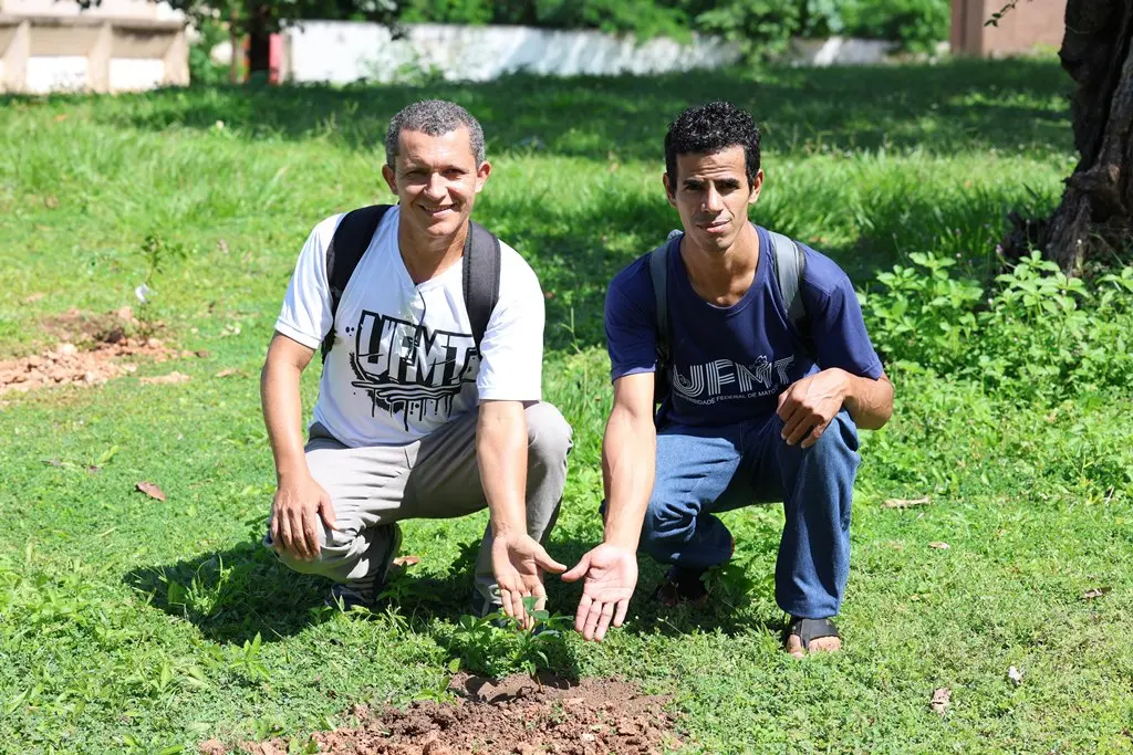Foto horizontal em plano aberto que mostra dois acadêmicos da UFMT agachados em volta de uma muda plantada e apontando para a planta. Eles usam camiseta com estampa da UFMT e estão em um campo aberto, todo gramado e com árvores ao fundo. 