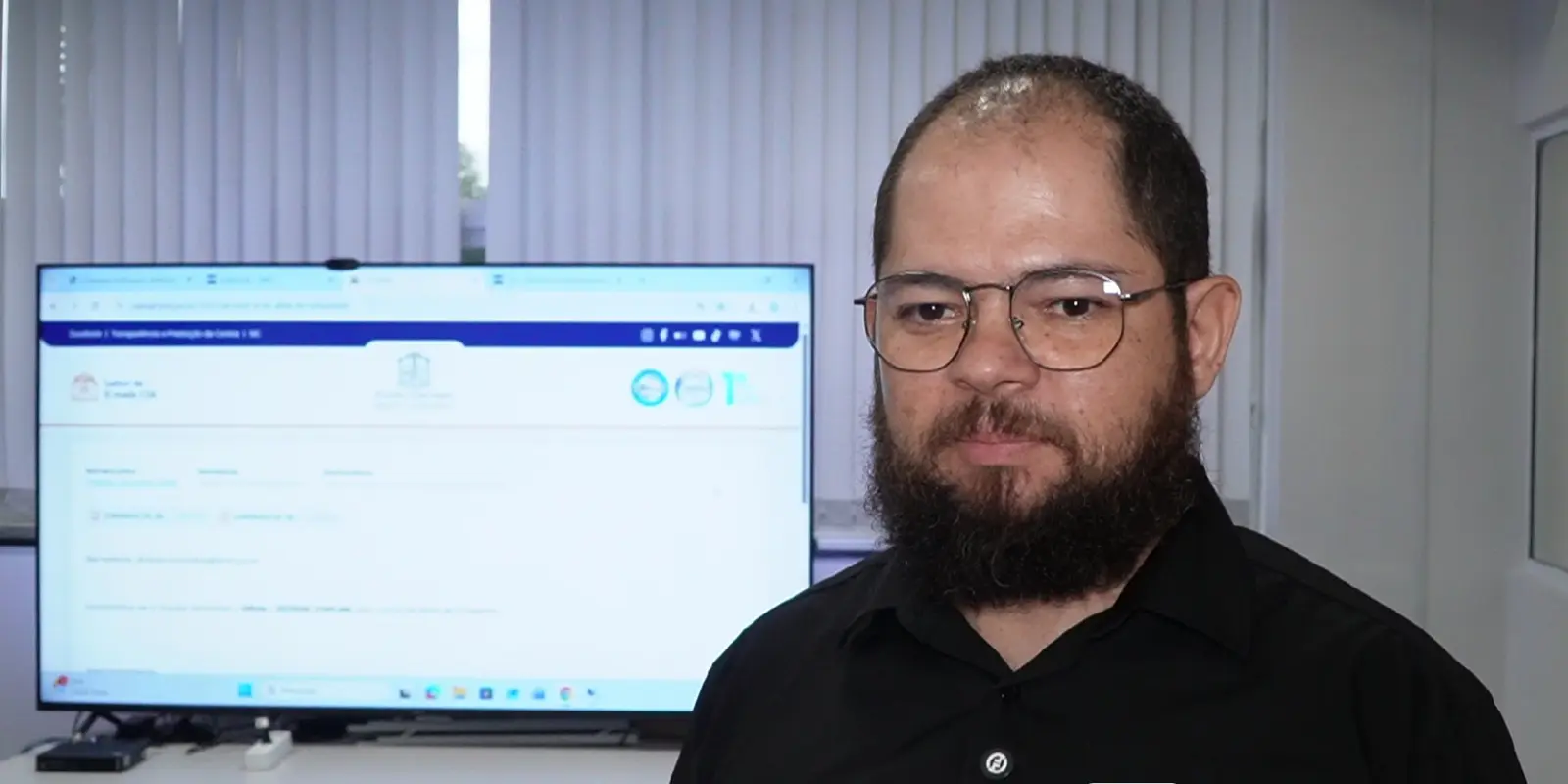 Um homem de cabelos escuros e barba preta, usa óculos de armação fina e veste camisa preta. Ao fundo, uma tela exibe um site institucional.