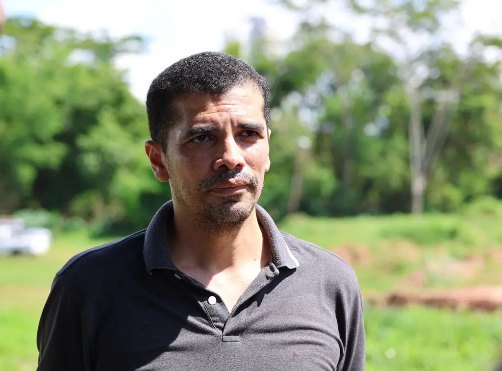 Foto horizontal em plano médio que mostra o professor Aldecy de Almeida Santos em pé, numa área de preservação permanente no campus da UFMT. Ele é um homem pardo, alto, magro, de olhos castanhos, cabelos castanhos, crespos e curtos, usando camiseta polo cinza. 
