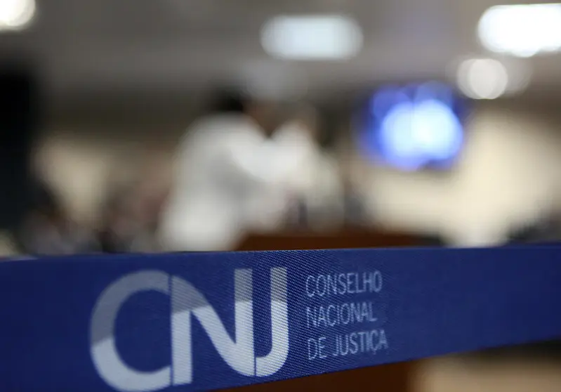Foto horizontal que mostra, em primeiro plano, uma faixa azul com a logomarca do Conselho Nacional de Justiça. O fundo da foto é desfocado, mas representa uma sala de audiência.