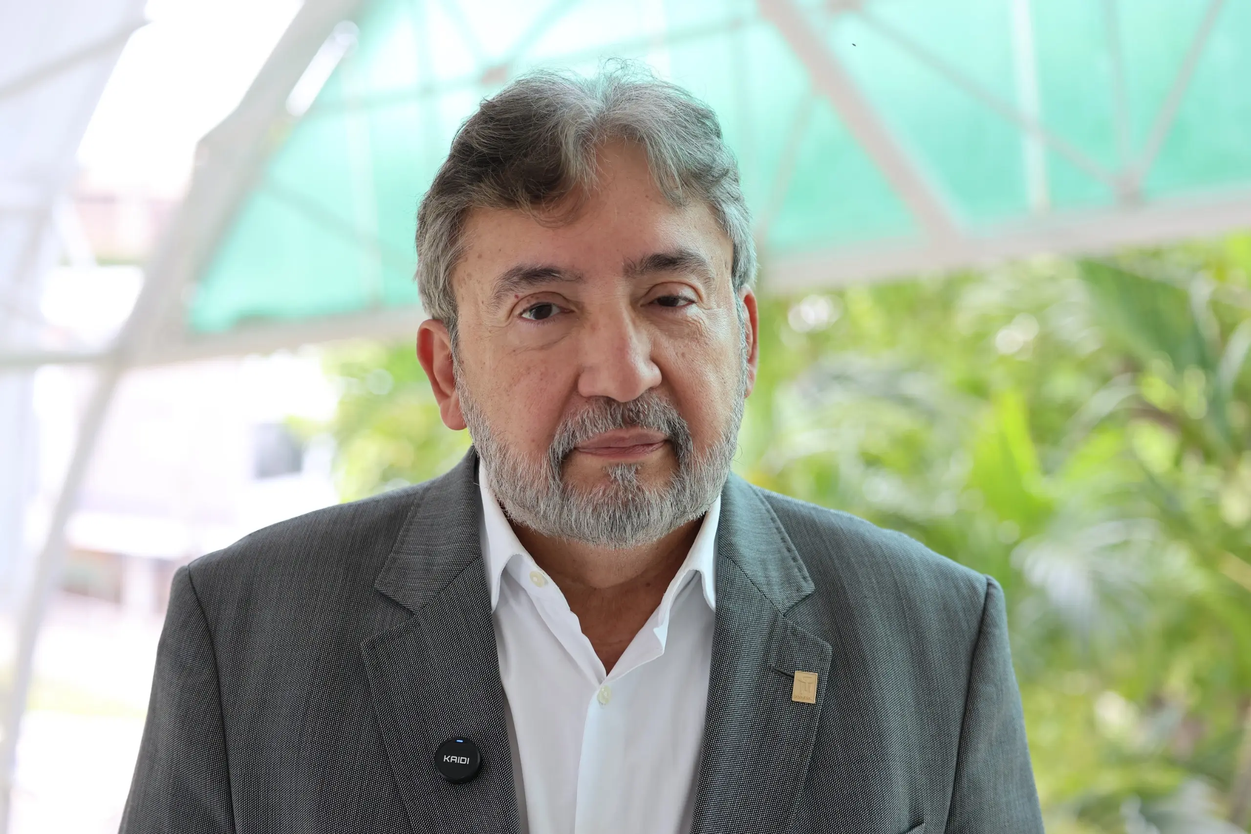 Imagem de um homem de barba e cabelos grisalhos, que veste um terno cinza e olha para a câmera. Ele está em um ambiente externo com bastante vegetação ao fundo.