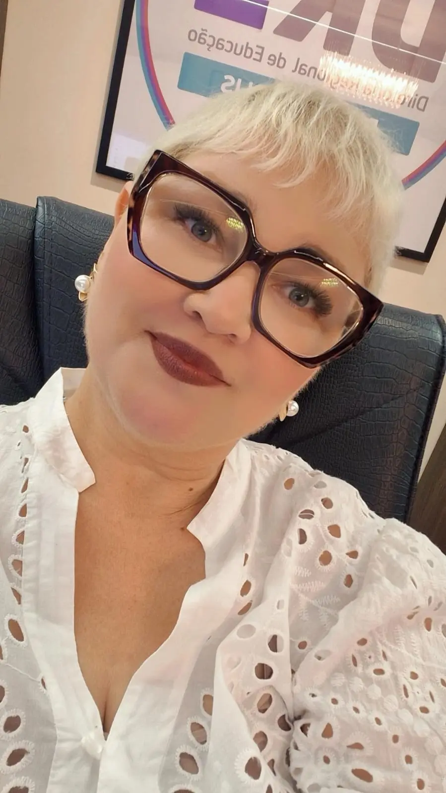 A imagem é um retrato de uma mulher de cabelos curtos e claros, usando óculos grandes e blusa branca rendada. Ela está sentada em uma cadeira de escritório, com expressão serena e ambiente profissional ao fundo.