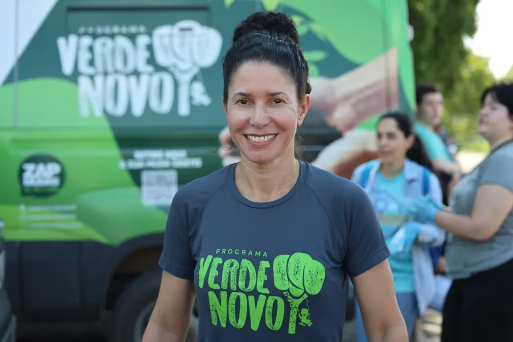 Foto horizontal em plano médio que mostra a engenheira florestal do programa Verde Novo, Rosiani Carnaíba, sorrindo para a foto, em frente à van do Verde Novo. Ela é uma mulher magra, de pele clara, olhos e cabelos escuros, usando camiseta com a logomarca do Verde Novo. 