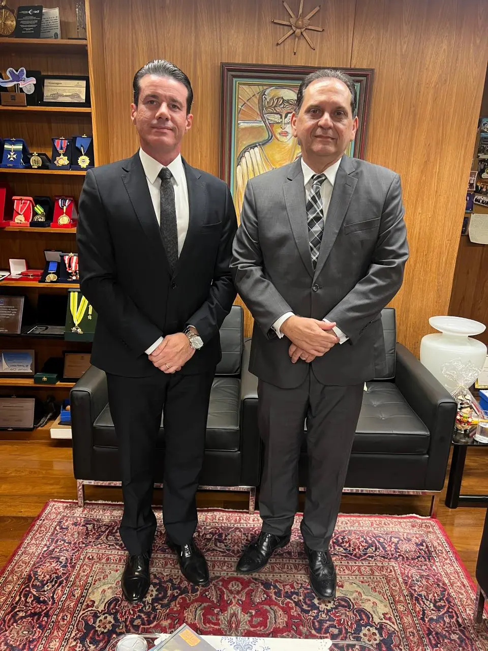 A foto mostra dois homens de terno escuro posando em um gabinete com paredes de madeira. Ao fundo há estantes com medalhas e homenagens, um quadro artístico e um sofá preto sobre tapete persa, em ambiente formal.