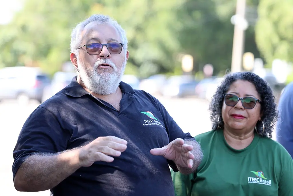 Foto horizontal em plano médio que mostra o presidente do Iteec Brasil, Valteir, em pé, falando e gesticulando, em uma área aberta desfocada. Ele é um senhor de pele branca, cabelos e barba brancos, usando camiseta polo preta com a logomarca do Iteec   Brasill bordada no peito e óculos de sol. 