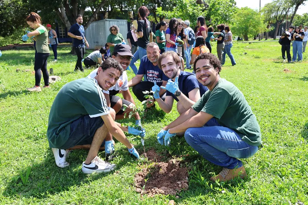 Foto horizontal em plano aberto que mostra, em primeiro plano, cinco rapazes agachados em volta de uma muda que estão plantando. Eles sorriem para a foto. Ao fundo, há uma movimentação de pessoas que também participam do plantio de mudas no campus da UFMT. 