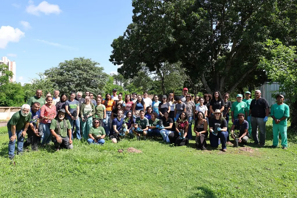Foto horizontal em plano aberto que mostra cerca de 50 pessoas reunidas, posando para a foto, algumas segurando mudas de plantas. Elas estão em uma área verde, com grama e árvores, no campus da UFMT. 