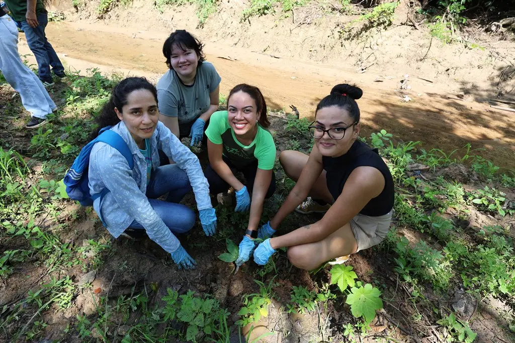 Foto horizontal em plano aberto que mostra quatro universitárias sorrindo para a foto, agachadas ao redor de uma muda, plantada na margem do afluente do Córrego do Barbado.