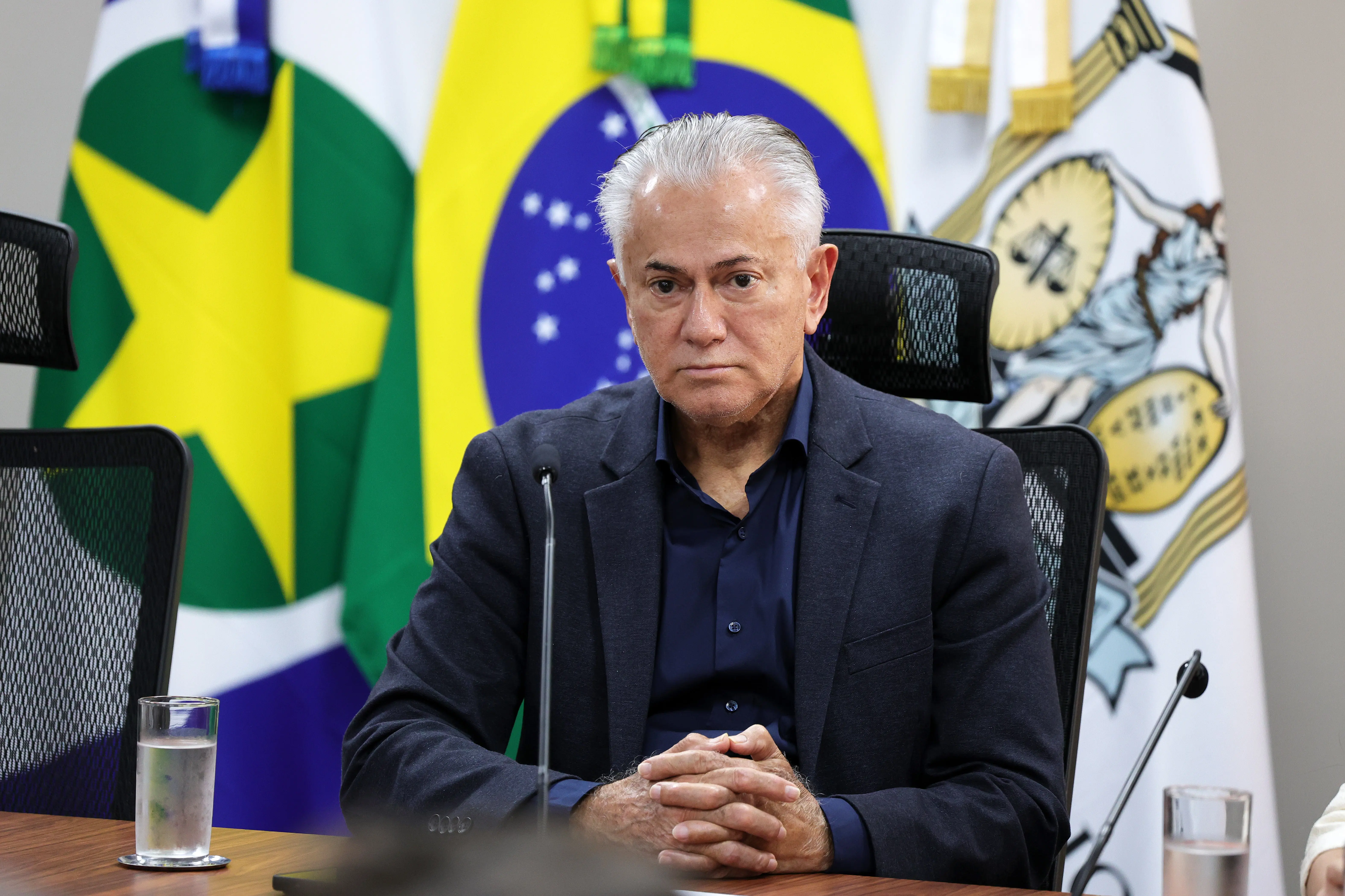 Homem de cabelos brancos, terno cinza-escuro e camisa azul-marinha, semblante sério, sentado à mesa com microfone, tendo ao fundo bandeiras do Brasil e de Mato Grosso.