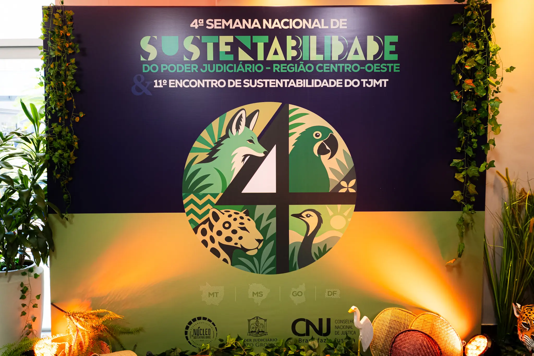 Painel do evento com o texto "4ª SEMANA NACIONAL DE SUSTENTABILIDADE DO PODER JUDICIÁRIO - REGIÃO CENTRO-OESTE". No centro, um círculo ilustra animais como raposa, tucano, onça e ema divididos pelo número 4.