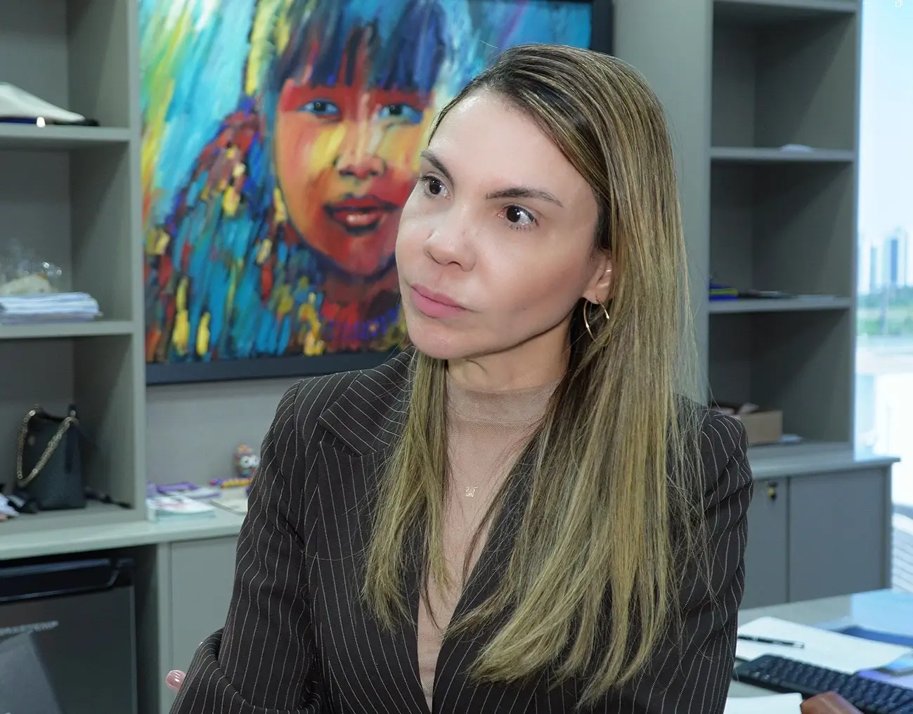 Foto horizontal que mostra a juíza Henriqueta Lima, do busto pra cima, olhando para o lado esquerdo. Ela é uma mulher de pele clara, cabelos longos, lisos e com mechas loiras, olhos castanhos, usando blusa marrom nude e blazer marrom escuro risca-de-giz. Atrás dela o ambiente é corporativo.