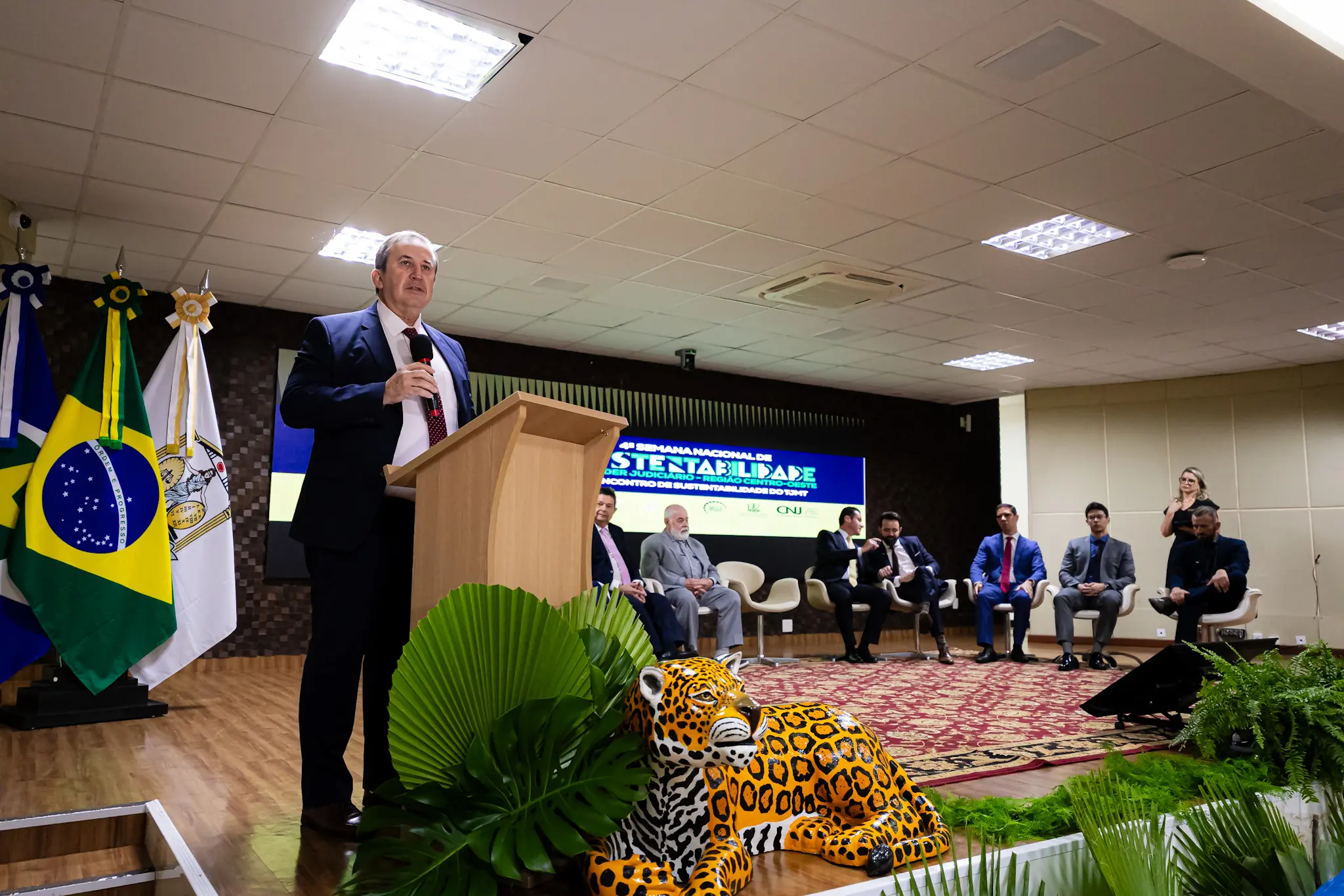 Visão ampla do palco do auditório. Um homem de terno discursa no púlpito, ao lado de bandeiras e de uma escultura de onça-pintada. No fundo, autoridade sentadas em cadeiras brancas diante de um painel do evento.