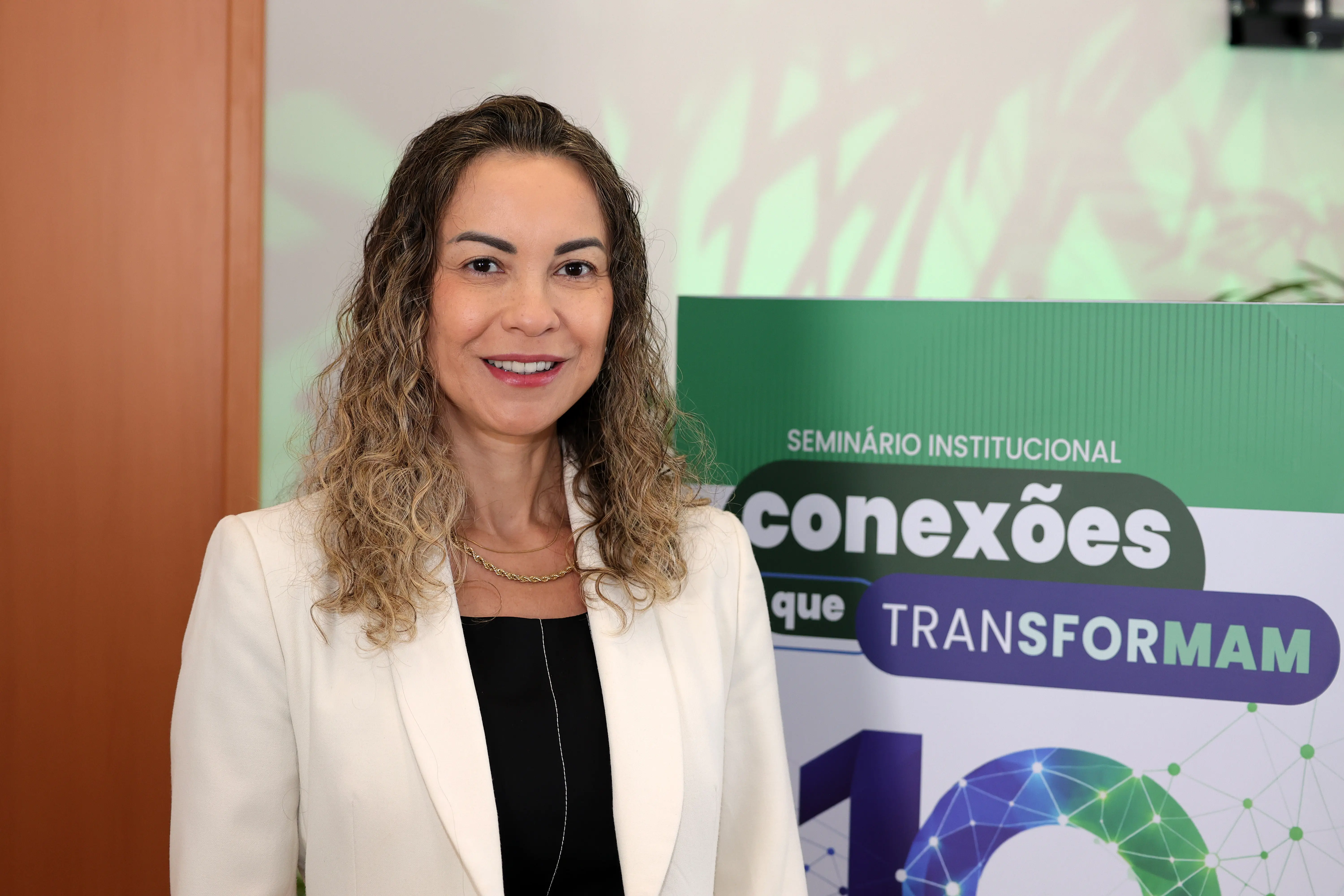 Mulher de cabelos castanhos ondulados até os ombros e blazer branco sobre blusa preta, sorrindo. Ao lado, um painel verde com o texto "SEMINÁRIO INSTITUCIONAL conexões que TRANSFORMAM 10".