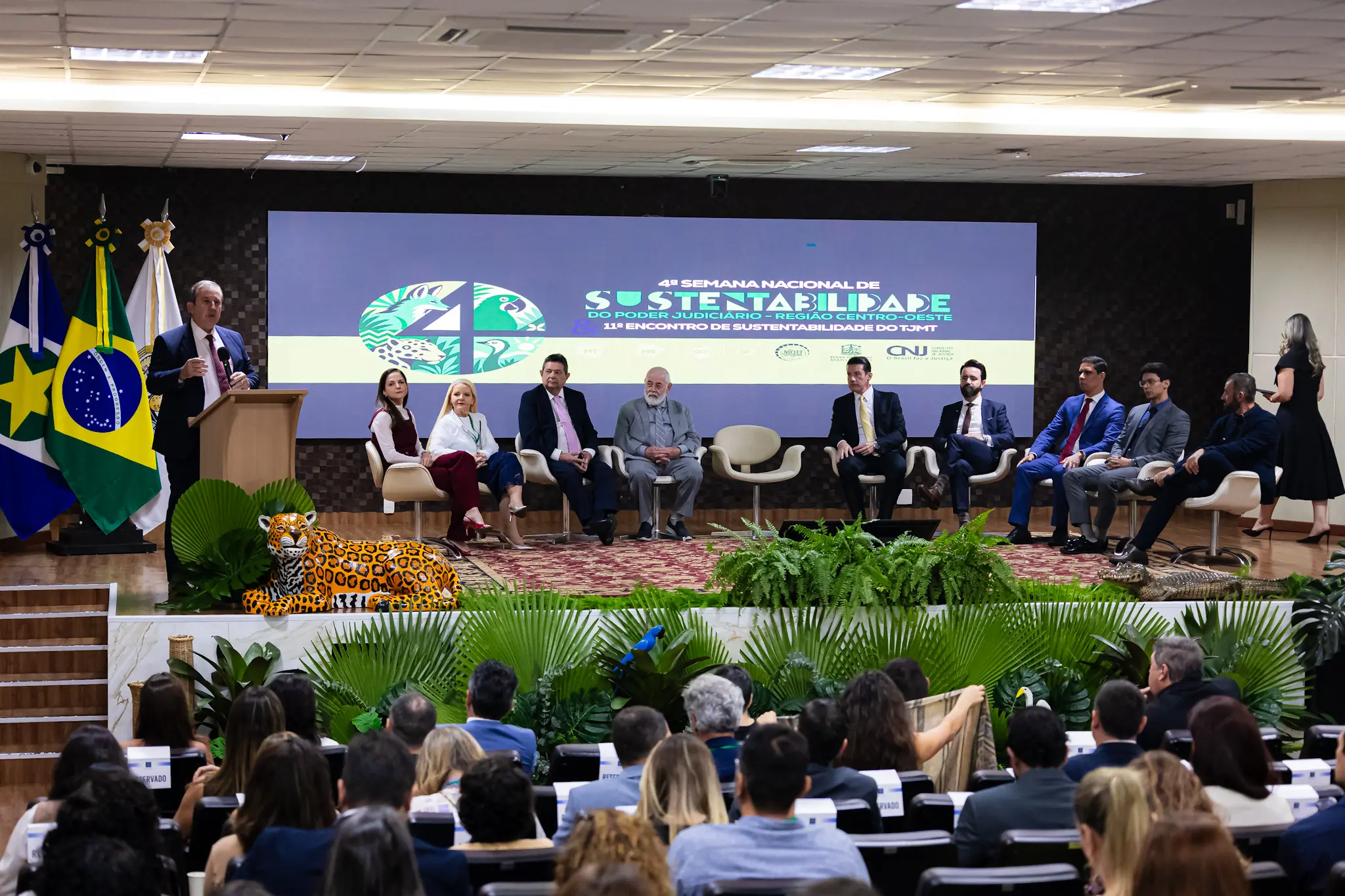 Plano geral do auditório visto de trás da plateia. No palco decorado com plantas e a escultura de uma onça, um homem discursa no púlpito enquanto outras autoridades permanecem sentadas ao fundo diante do painel do evento.
