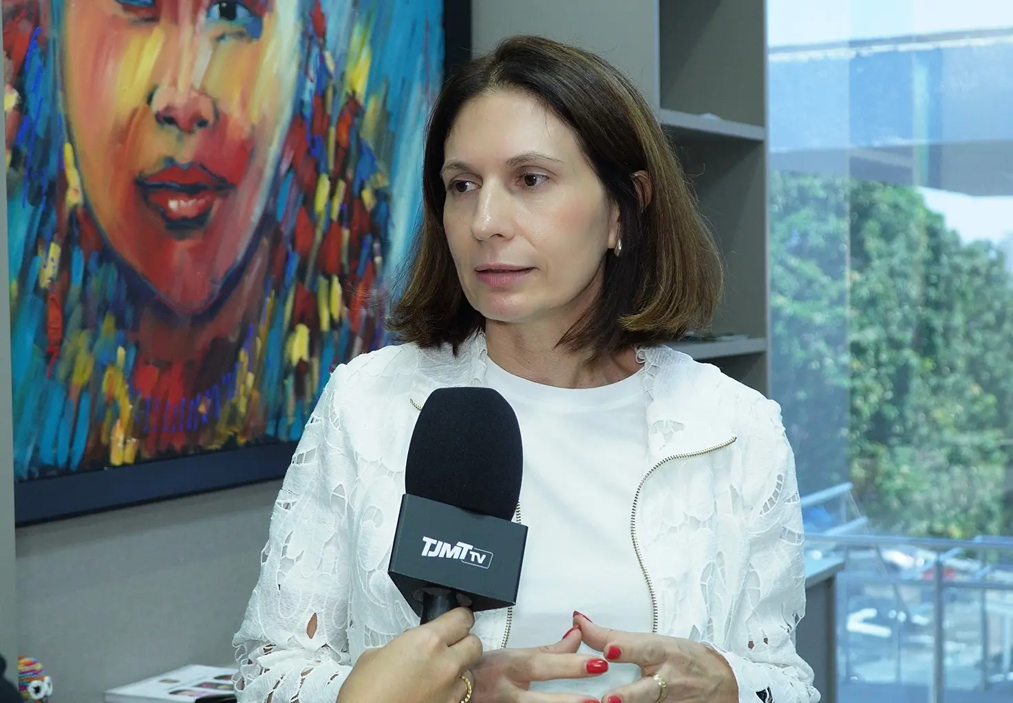 Foto horizontal que mostra Patrícia Cocatto, do busto pra cima, durante entrevista à TJMT TV. Ela é uma mulher branca, de cabelos castanhos, lisos em corte chanel, usando blusa branca com blazer branco em renda floral. Atrás dela, aparece um quadro que retrata o rosto de uma menina indígena. 