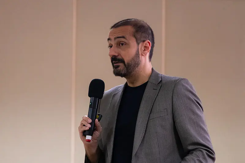 Foto horizontal que mostra o advogado e palestrante Fabrício Soler, do busto pra cima, falando ao microfone, no palco. Ele é um homem branco, alto, magro, calvo, de barba e olhos pretos, usando camiseta preta e blazer cinza.