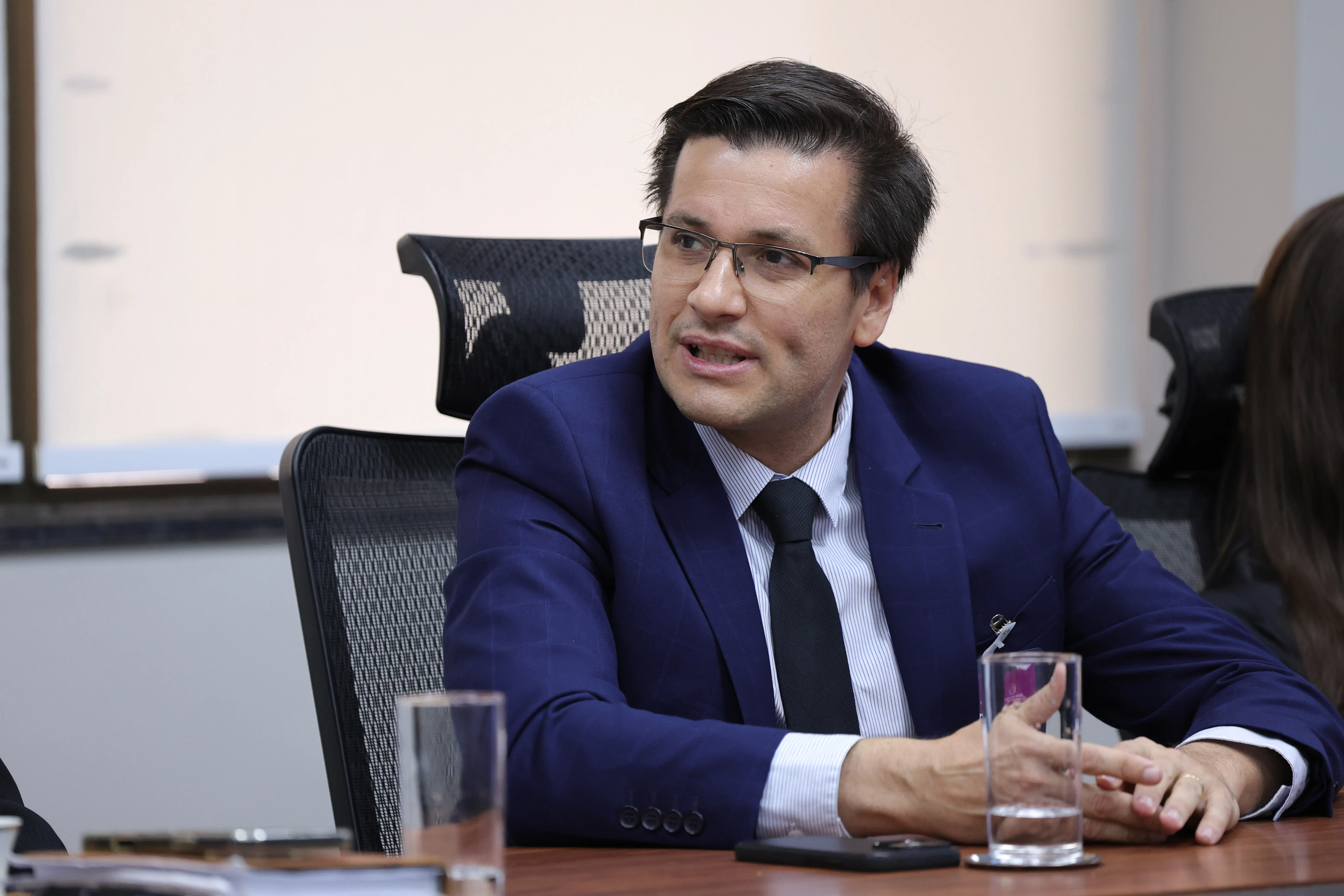 Homem de cabelos escuros, óculos de grau e terno azul-marinho, camisa listrada e gravata preta, gesticula durante reunião em sala do TJMT, com copos d'água à mesa.