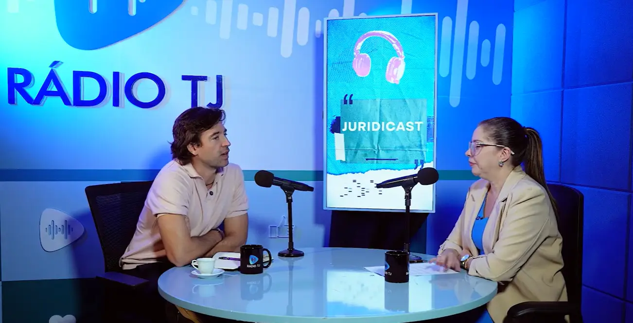 Em estúdio iluminado em tons azuis, um homem de camisa polo rosa e uma mulher de blazer bege com blusa azul conversam sentados a uma mesa redonda. À frente de cada um há microfones e canecas. Ao fundo, um painel vertical exibe a arte do "JURIDICAST" e a parede traz o letreiro "RÁDIO TJ".