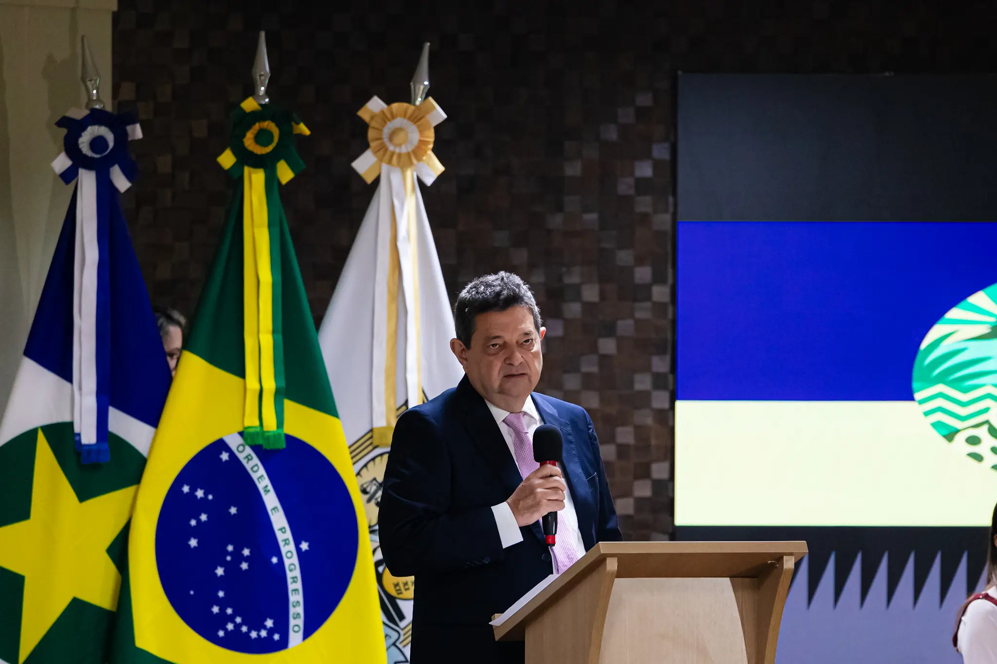 Homem de terno escuro e gravata rosa claro discursa em um púlpito de madeira segurando um microfone. À esquerda, três bandeiras hasteadas: de Mato Grosso, do Brasil e uma branca. Ao fundo, há um painel colorido.