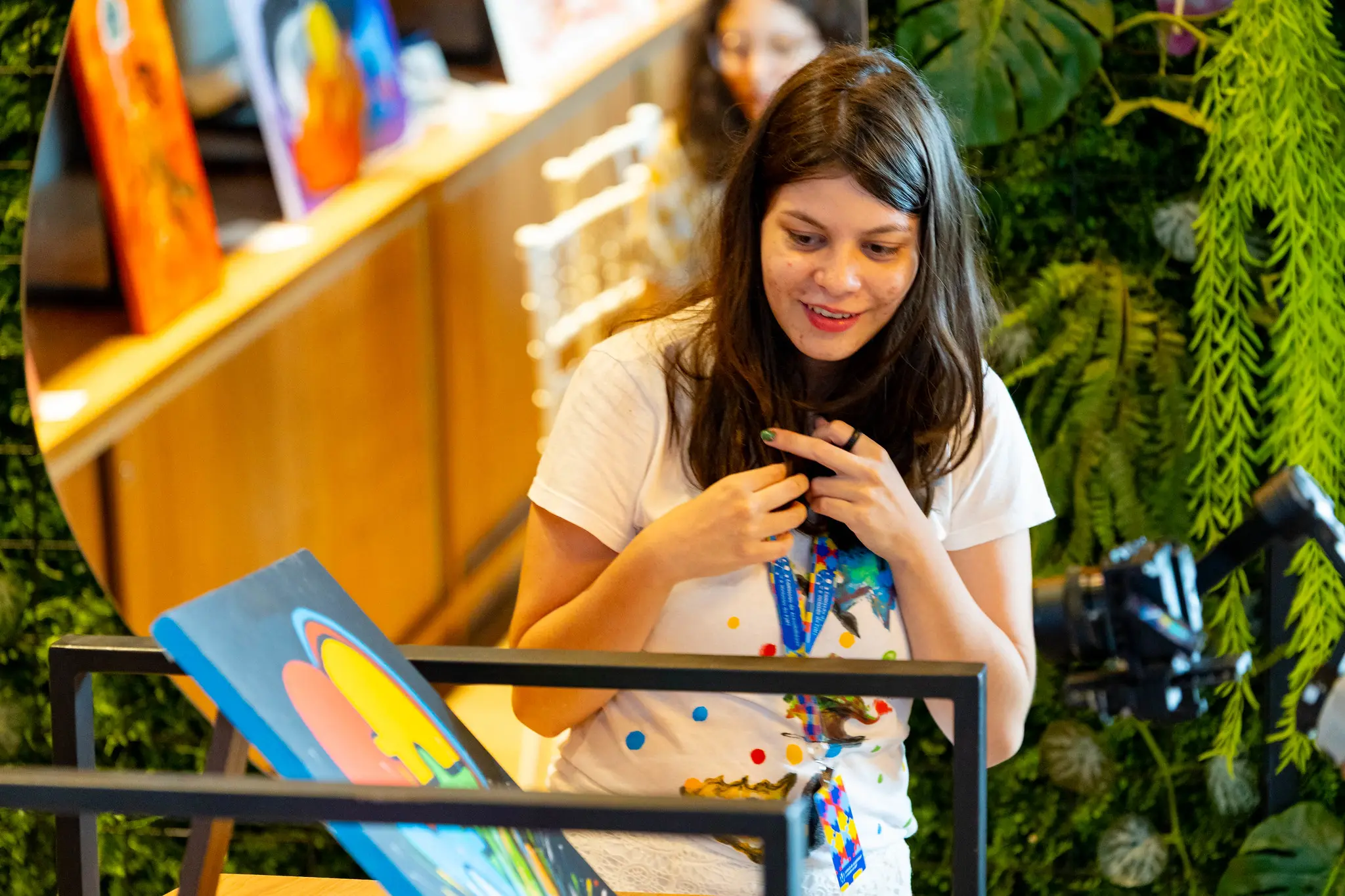 Maria Clara observa uma de suas telas expostas, sorrindo e com expressão concentrada. Ela está em seu estande no TJMT Inclusivo, rodeada por plantas decorativas, prateleiras e suas obras coloridas, criando clima artístico e acolhedor.