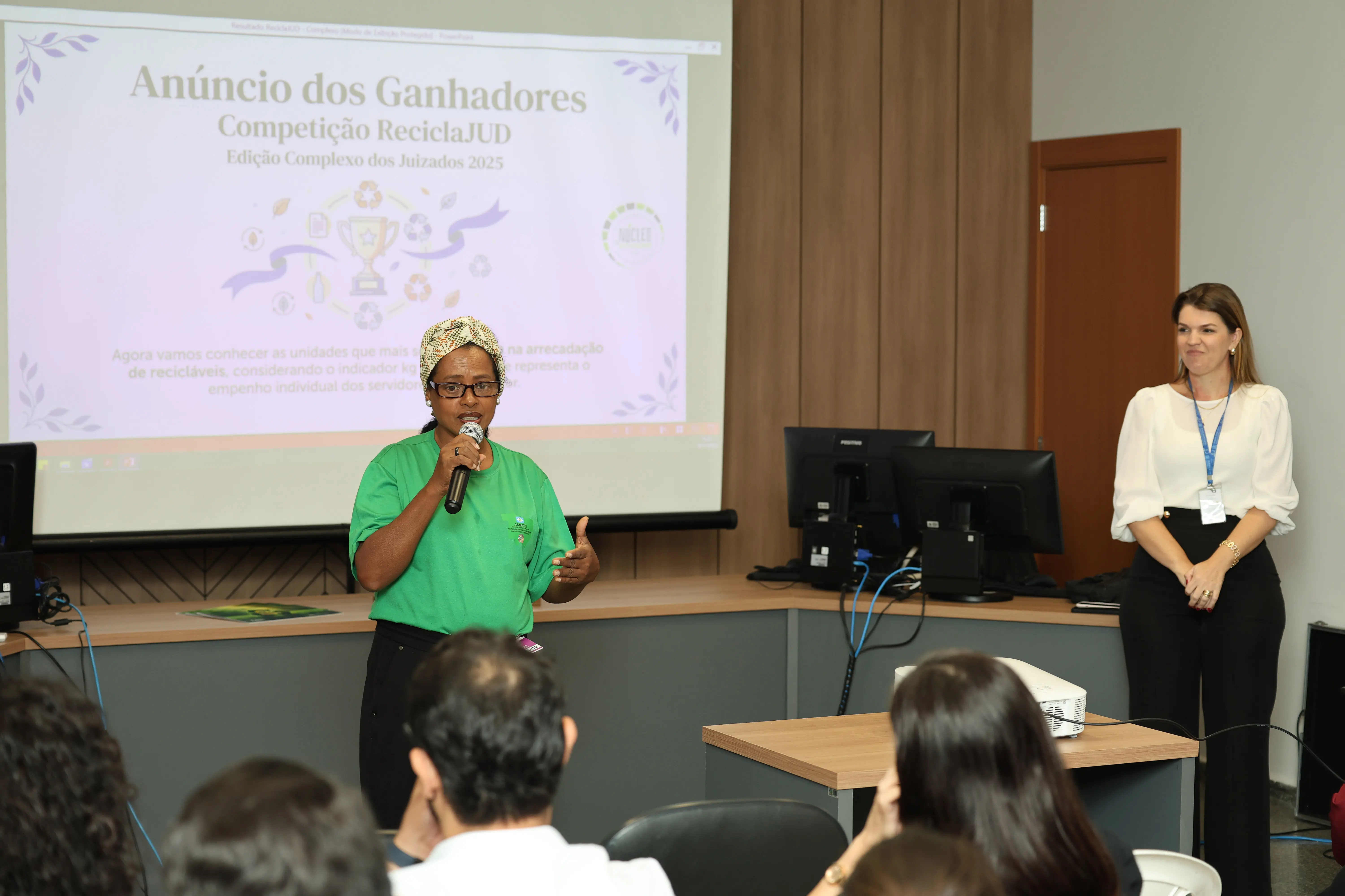 Mulher de camisa verde e turbante fala ao público com microfone, diante de telão do ReciclaJud. À direita, servidora com crachá observa. A sala tem computadores e projetor, ambiente de treinamento.