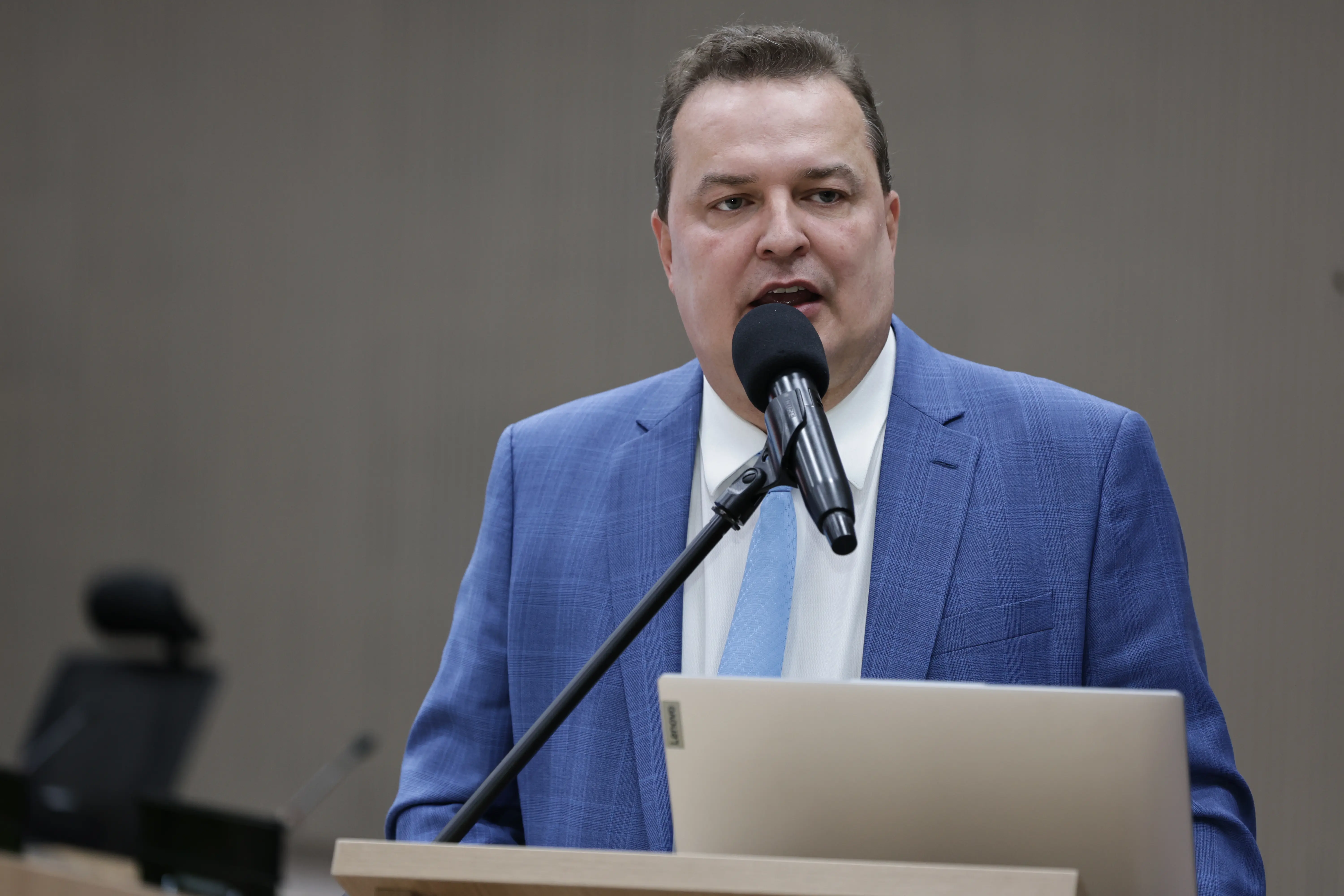 Em um auditório institucional, um homem de terno azul-claro e gravata em tom pastel discursa ao microfone, posicionado em um púlpito com notebook. Seu semblante é sério e o fundo neutro reforça o ambiente formal da solenidade.