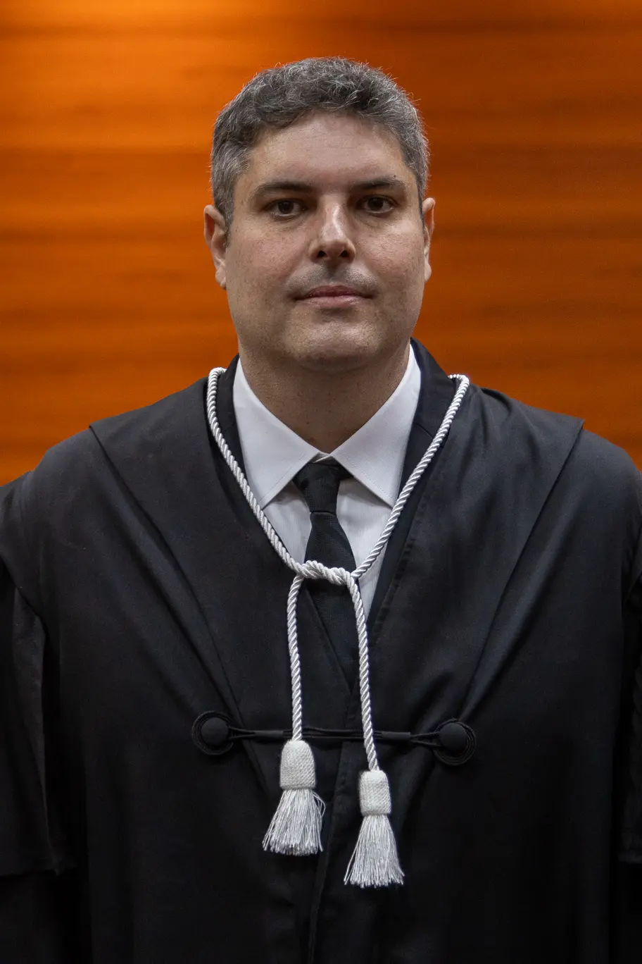 Magistrado posa em ambiente interno com parede de madeira ao fundo. Veste toga preta com cordão branco cerimonial e gravata escura, olhando diretamente para a câmera com expressão formal.