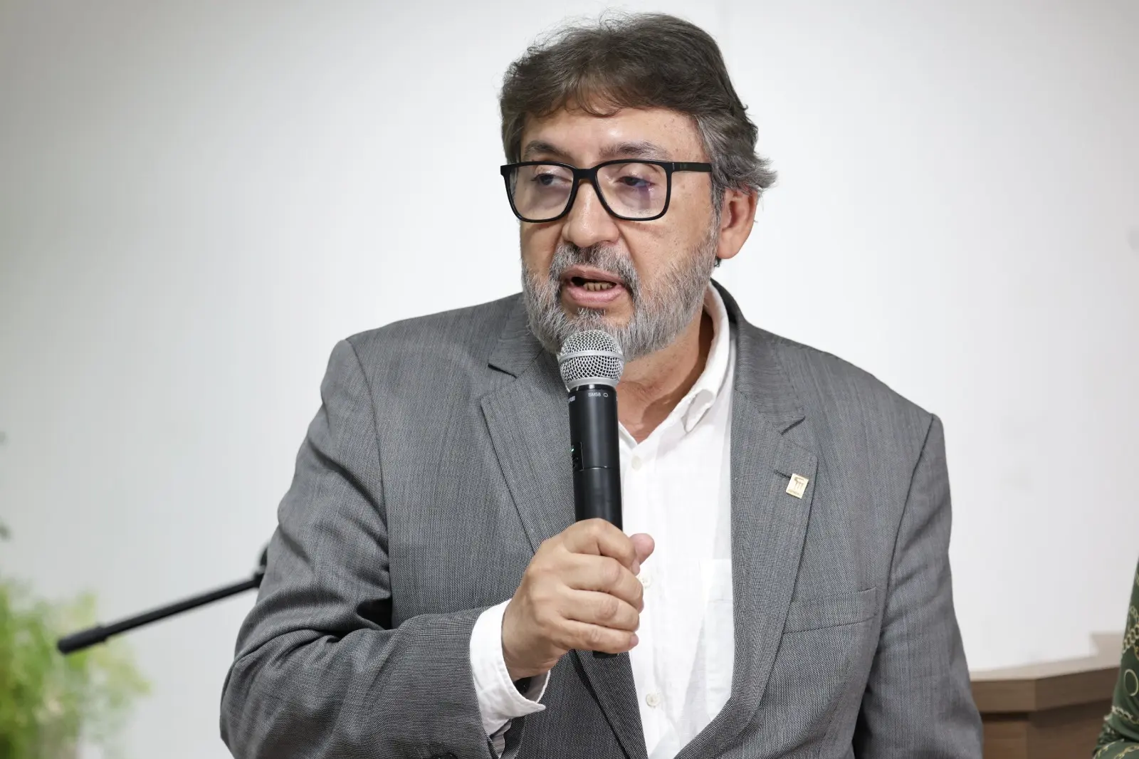 Foto do des. Márcio Vidal. Um homem de meia-idade com barba grisalha e óculos de armação preta está em close-up, falando em um microfone. Ele veste um blazer cinza risca de giz sobre uma camisa social branca, com um pin dourado na lapela.