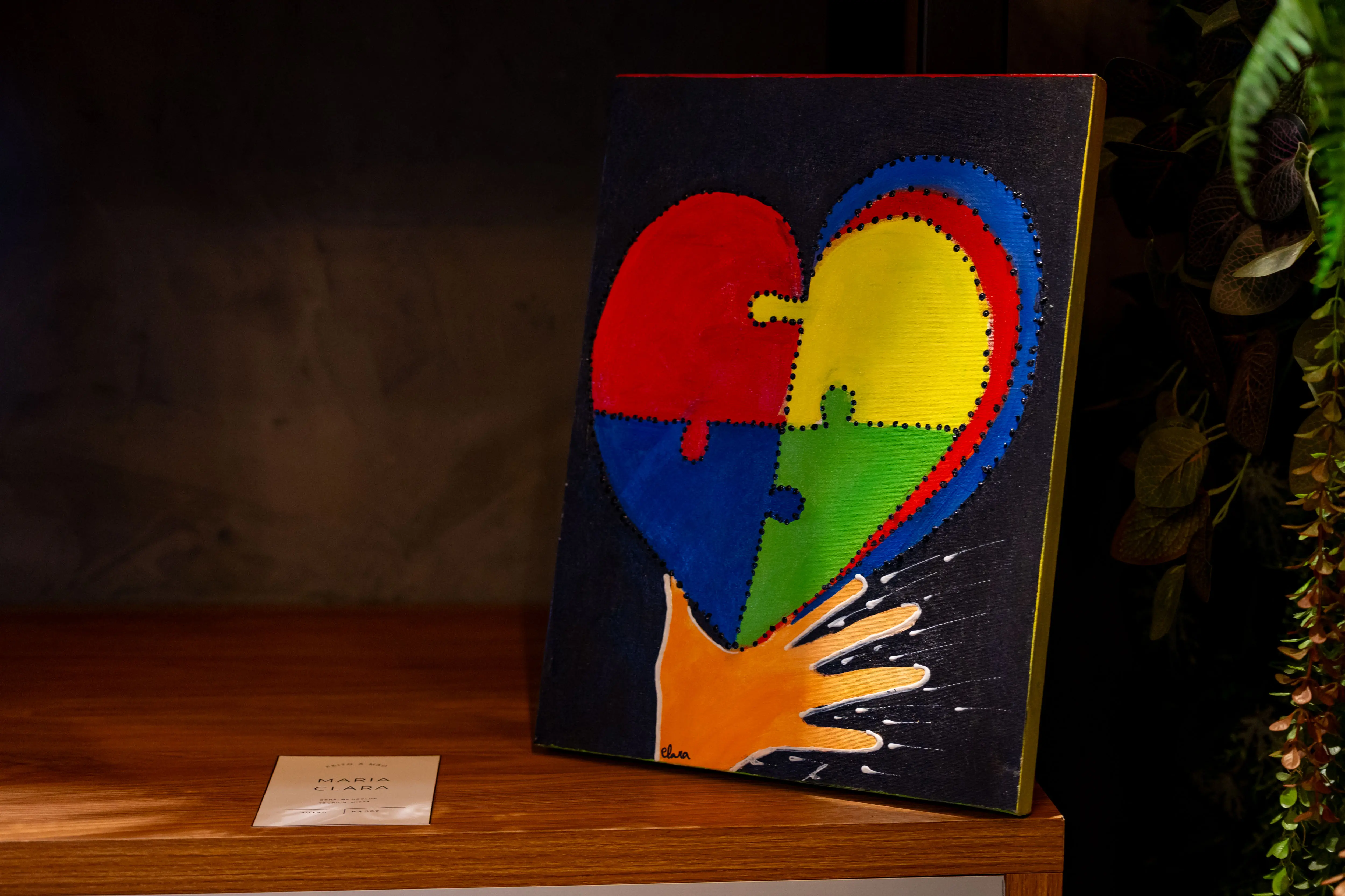 Uma tela com coração estilizado em quatro cores simbolizando o autismo ocupa o centro da foto. O quadro, exposto no estande de Maria Clara, está apoiado em prateleira de madeira em espaço decorado com plantas e iluminação suave.