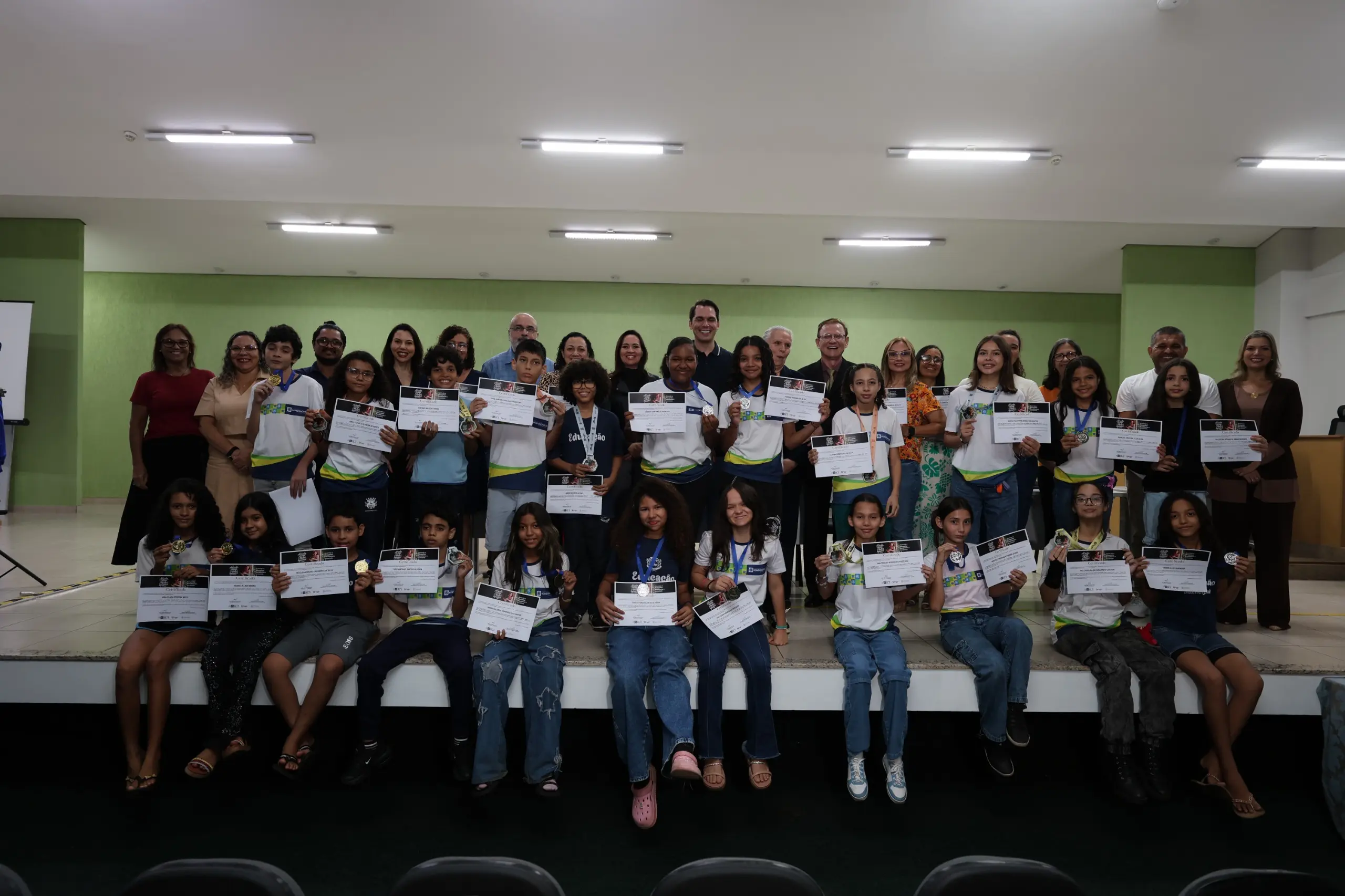 Uma foto de grupo em um palco, com mais de 30 pessoas, incluindo adultos e cerca de 18 crianças e adolescentes. As crianças estão segurando certificados e medalhas, sentadas ou em pé na frente, indicando uma premiação