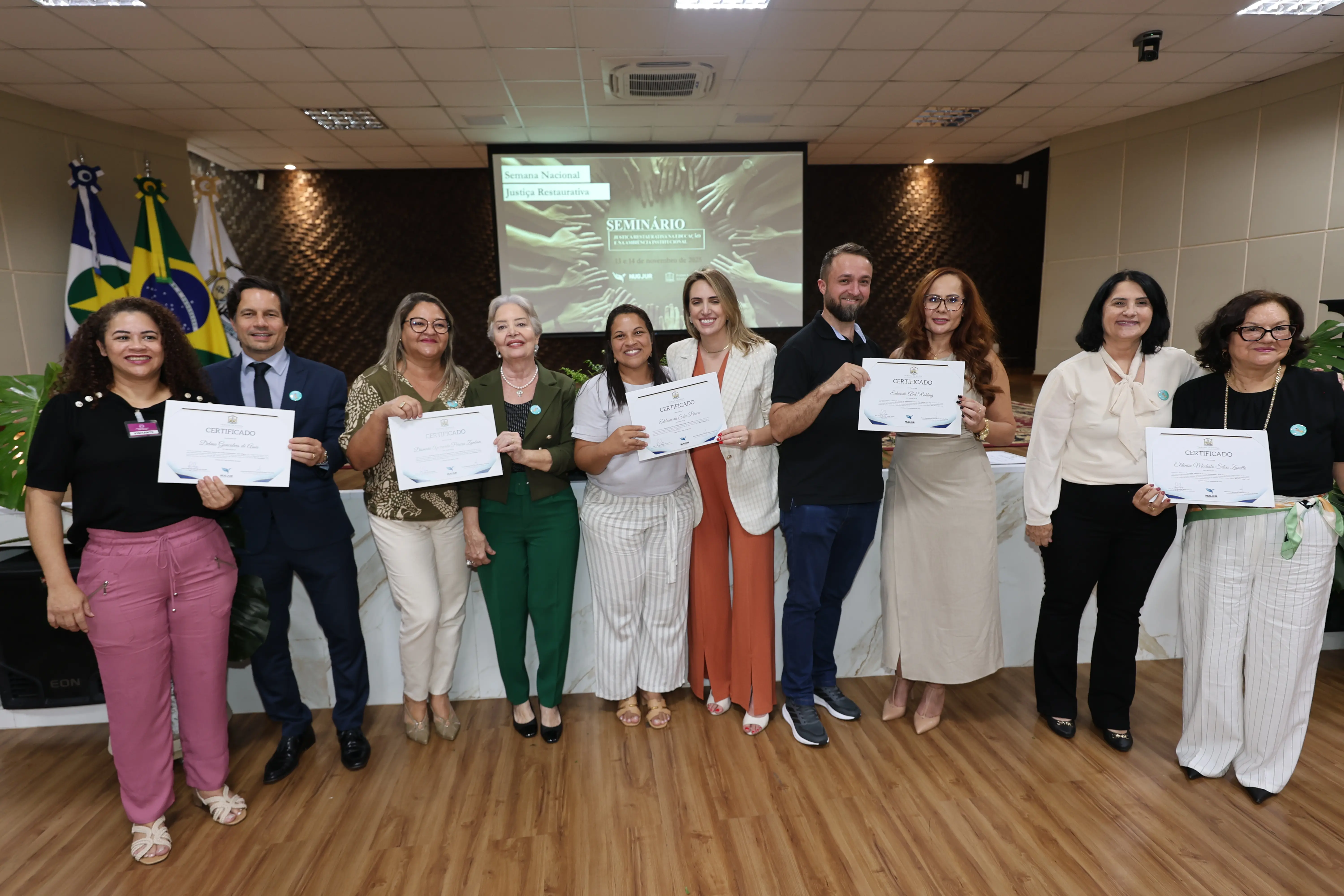 Grupo de nove pessoas (três homens e seis mulheres) em pé, lado a lado, sorrindo e segurando certificados de um seminário. Bandeiras estão visíveis no canto esquerdo.