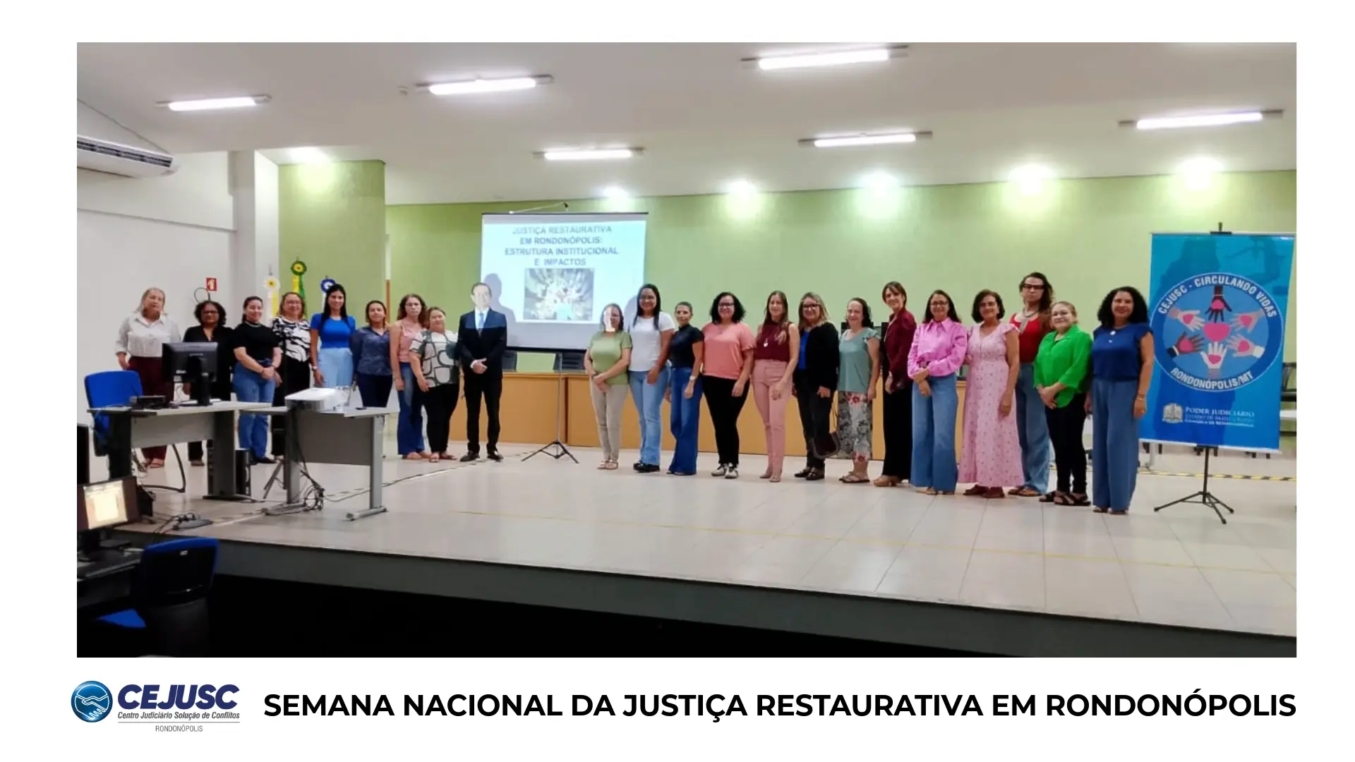 Grande grupo de servidoras e servidores posa em auditório iluminado, diante de telão com tema da Justiça Restaurativa. Estão alinhados em semicírculo, sorrindo, ao lado do banner do Cejusc de Rondonópolis.