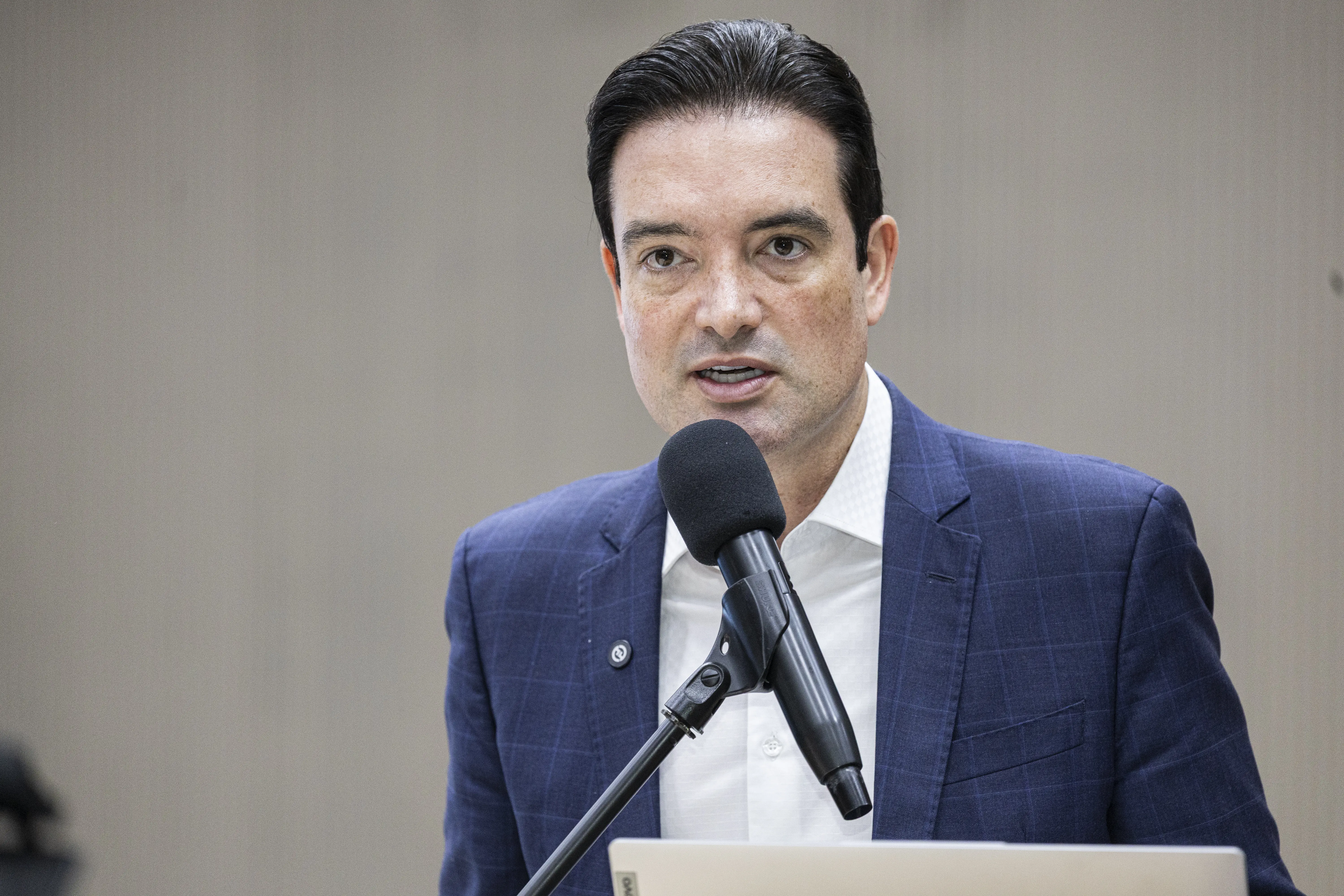 Um homem de terno azul-escuro e camisa branca fala ao microfone em um auditório oficial. Com expressão firme, ele está ao púlpito, diante de fundo claro e neutro, compondo a atmosfera formal do evento institucional.