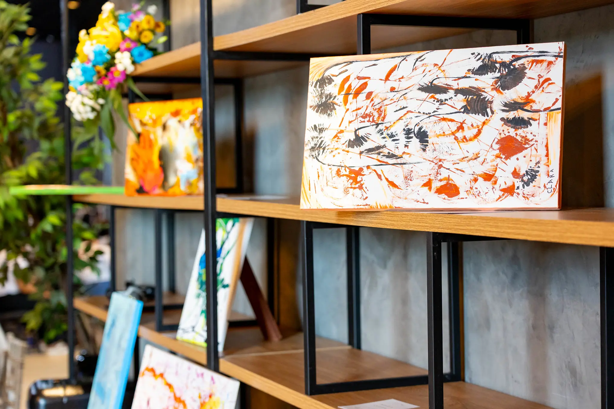 Prateleiras de madeira exibem diversas telas pintadas por Maria Clara, com cores vibrantes e traços abstratos. O local é o estande no TJMT Inclusivo, organizado com arranjos florais e peças de arte distribuídas pelo ambiente.