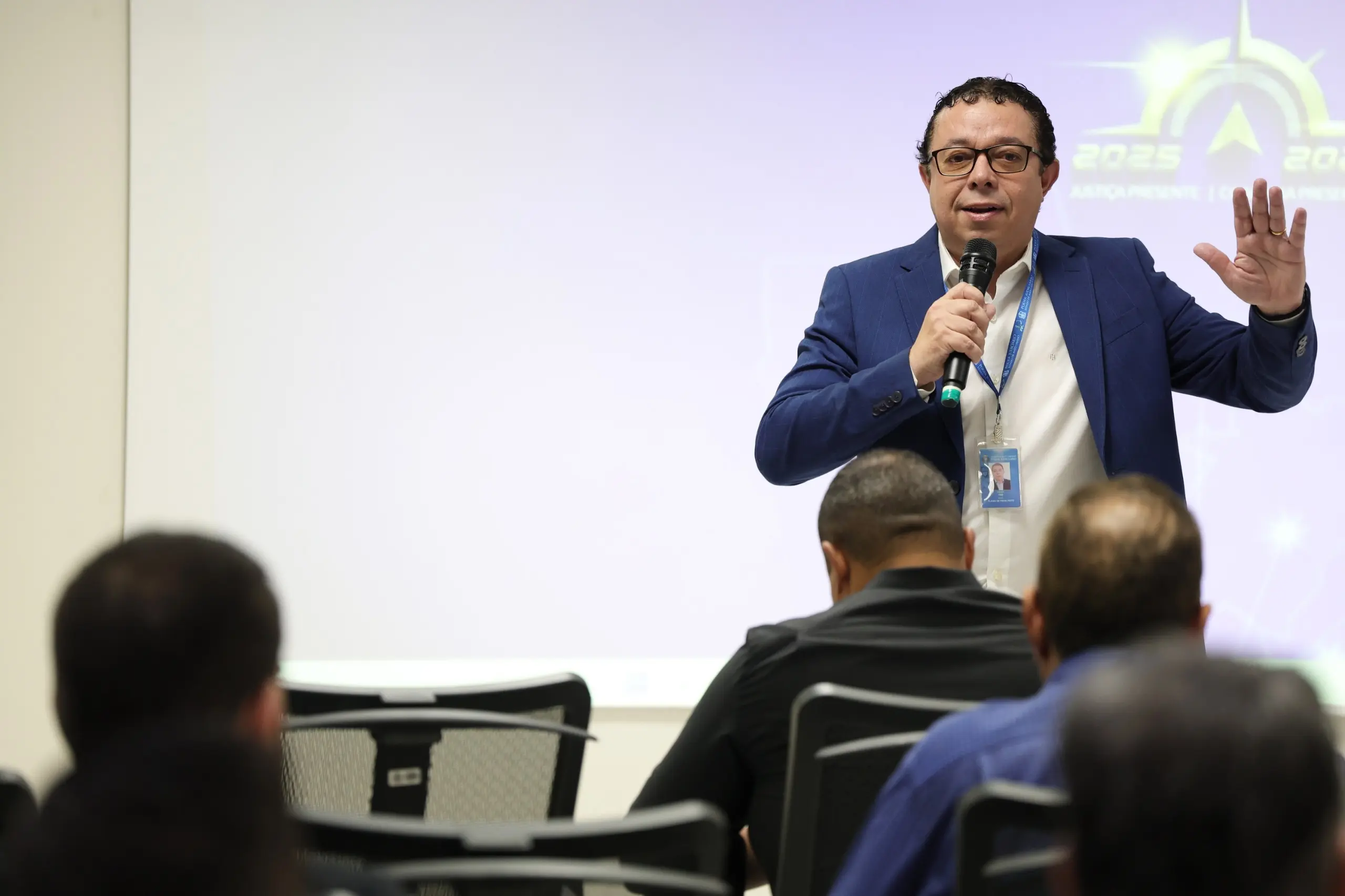 Homem de terno azul fala ao microfone diante de participantes sentados em uma sala. Ao fundo, uma projeção com elementos gráficos do evento.